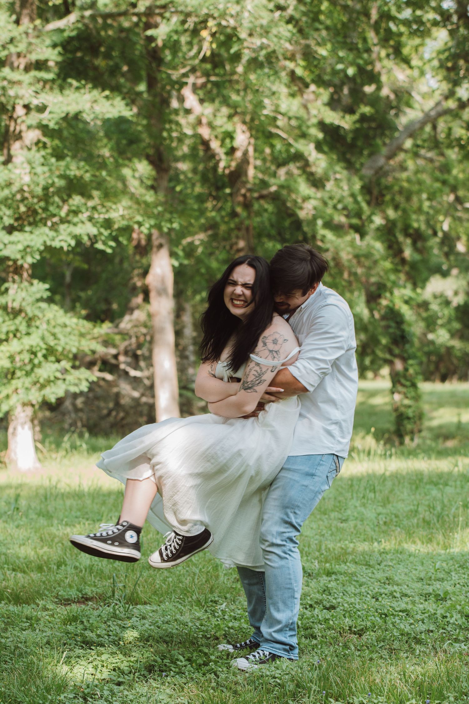 A playful couple embraces while wearing casual clothes in a sunny park with green trees in the background.
