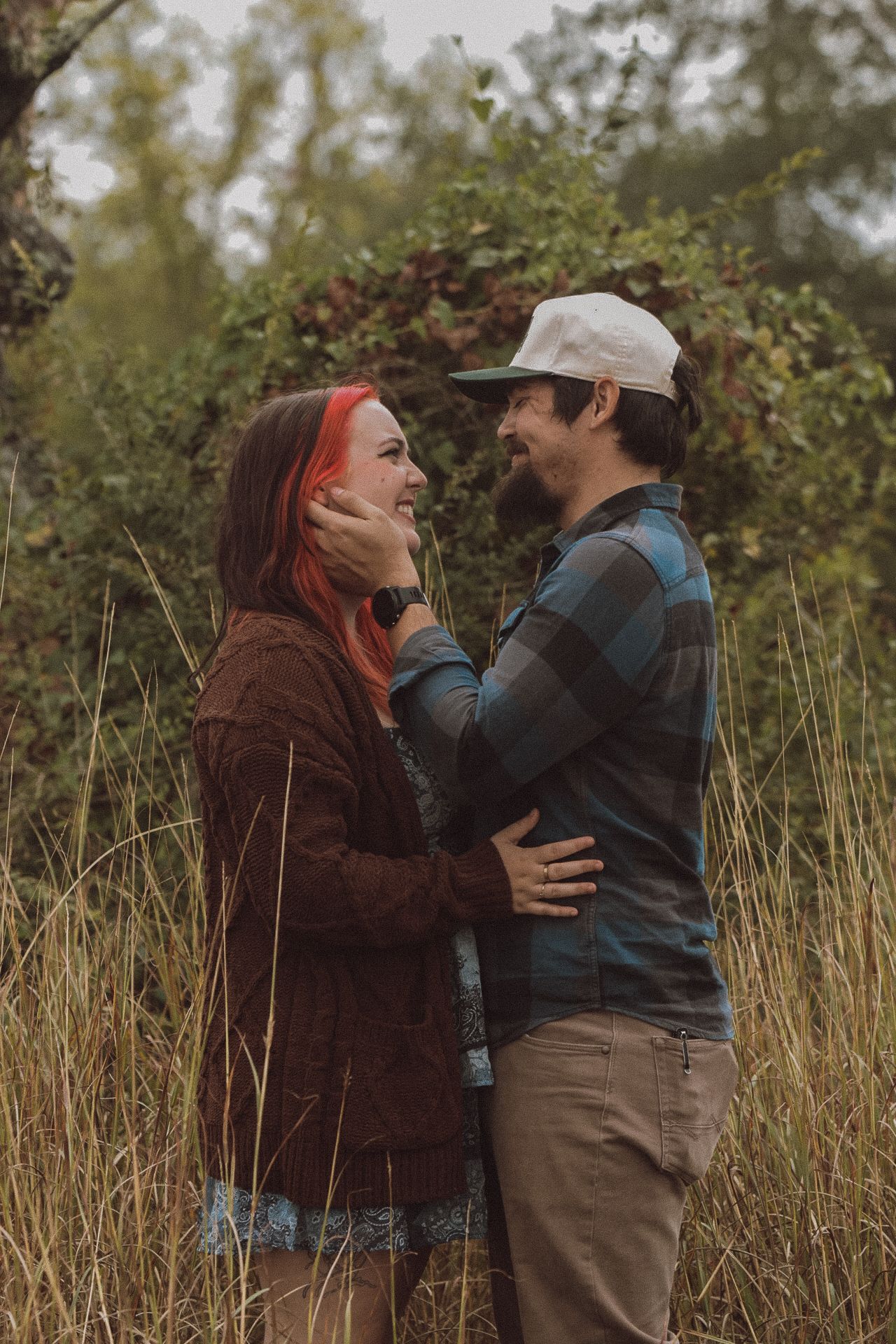 Couple embracing in tall grass field, woman with red hair and man in plaid shirt and cap, surrounded by lush greenery.
