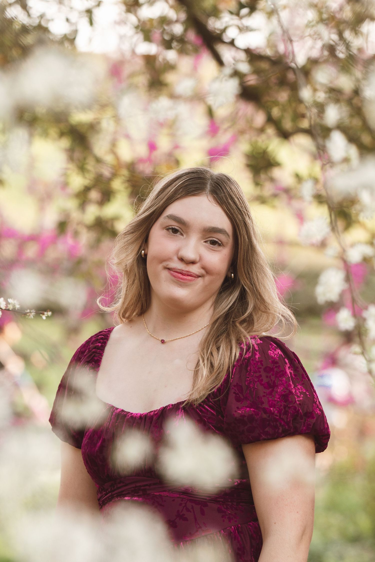 Outdoor portrait with burgundy velvet dress against soft pink spring blossoms and bokeh background.