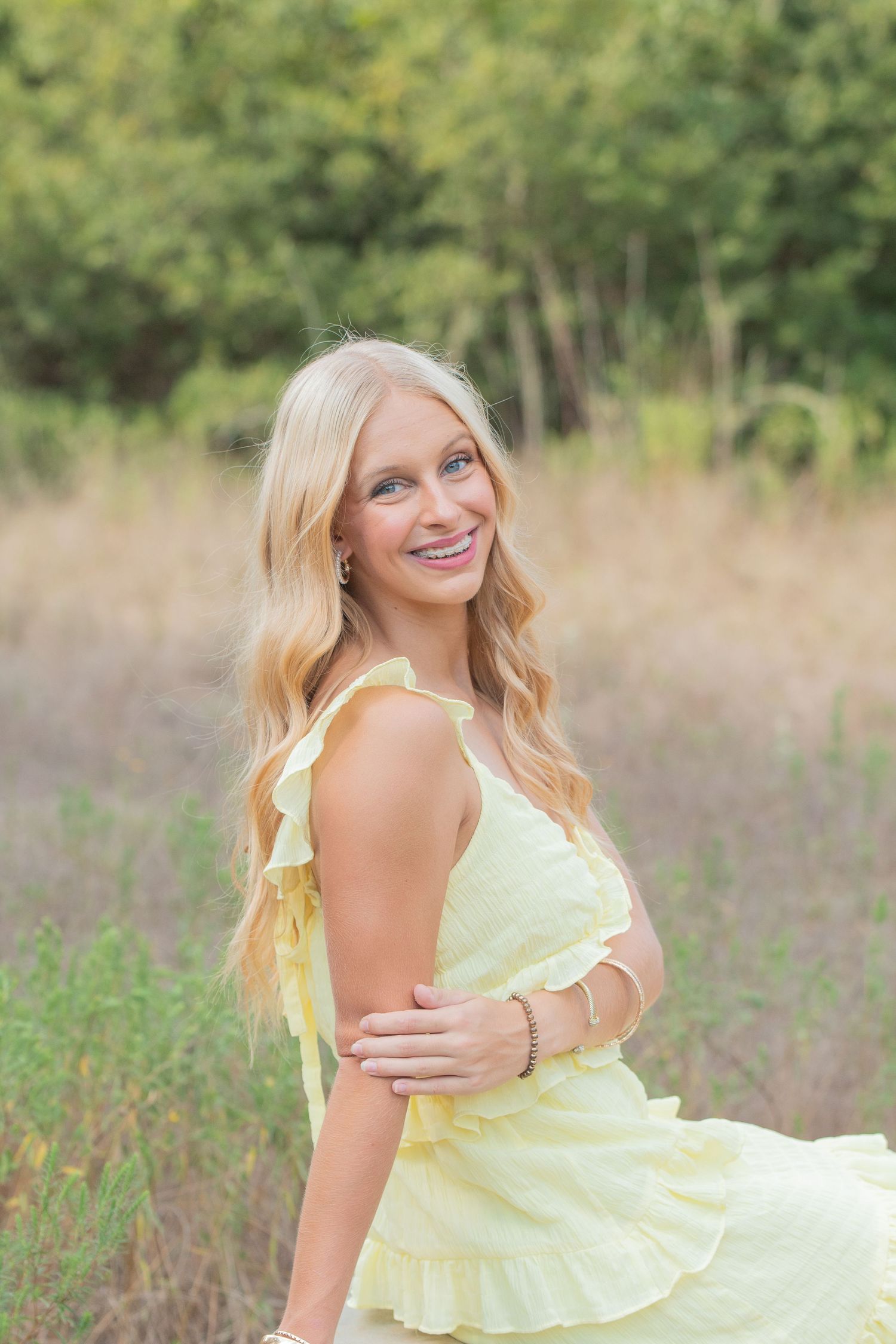 A woman in a flowing yellow sundress poses gracefully in a meadow with trees in the background.