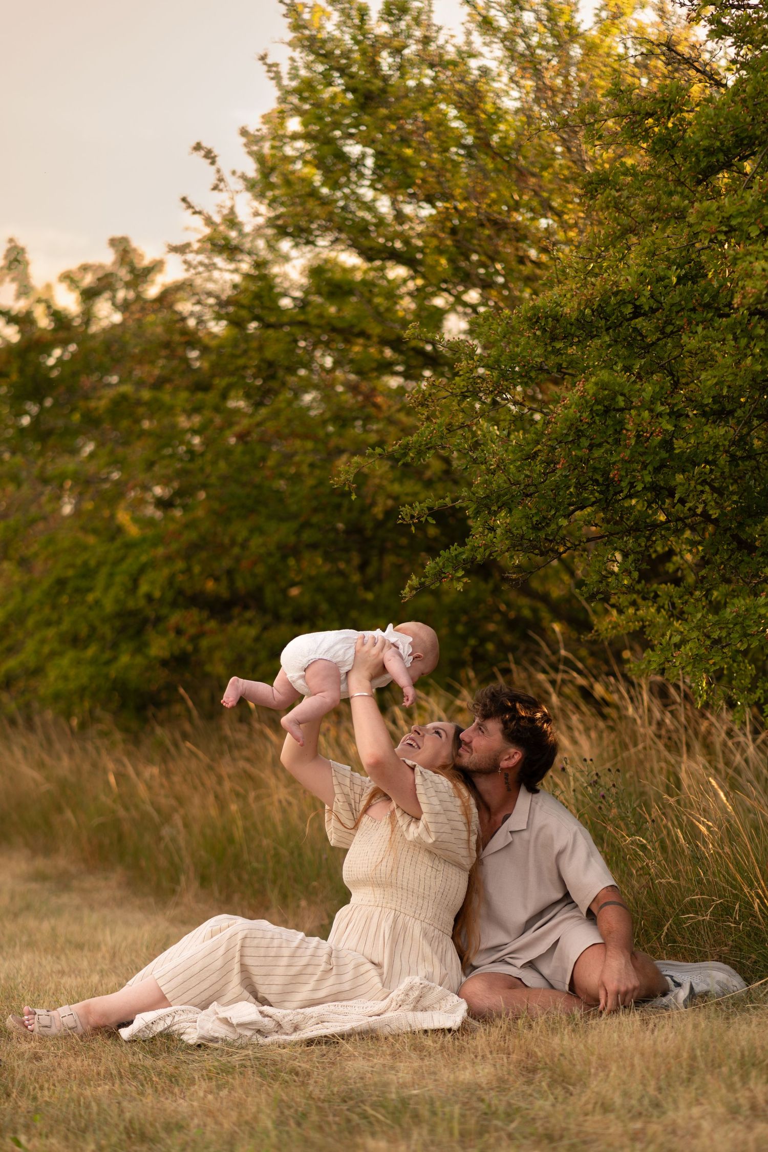 A couple sits in a golden field, lovingly lifting their baby in the air, surrounded by lush green trees at sunset.