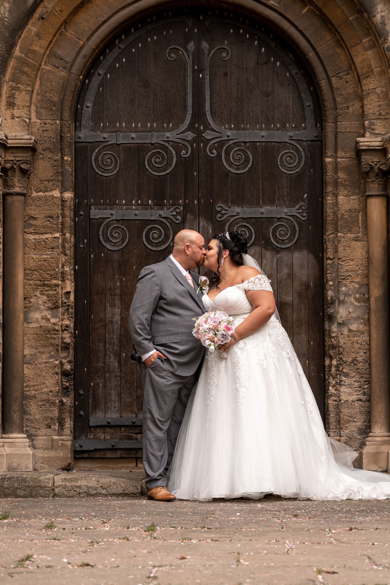 A romantic wedding couple shares an intimate moment against a historic stone church with ornate iron doors.