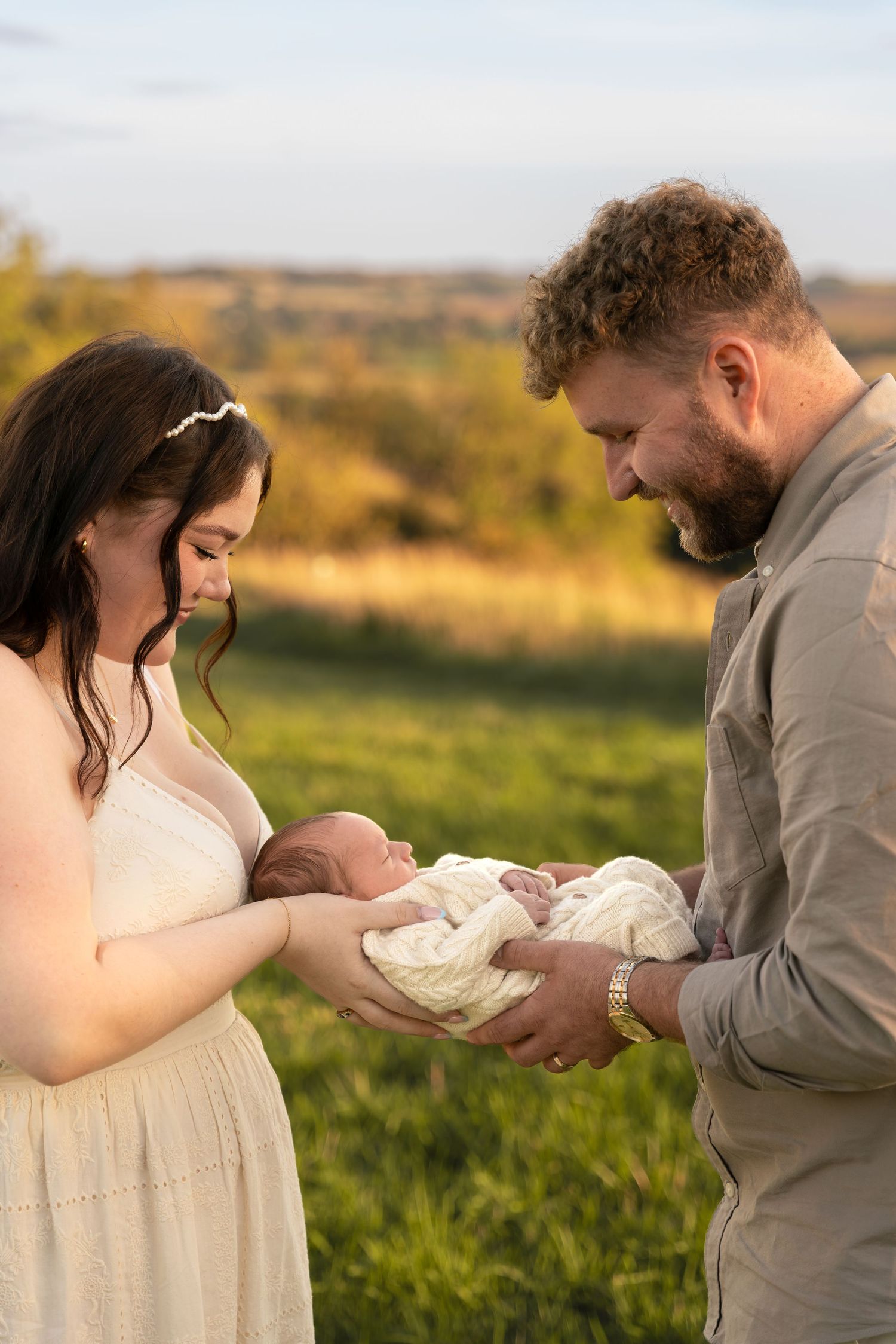 A couple tenderly holds their newborn baby outdoors in a golden field during a family portrait session.
