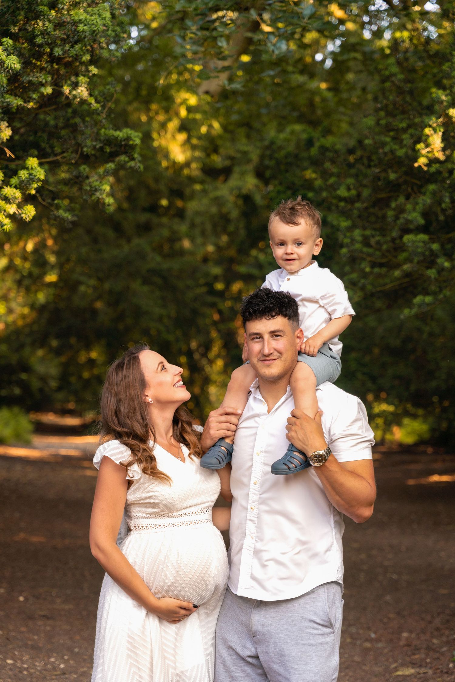 Pregnant woman in white dress with husband carrying toddler on shoulders, smiling outdoors during a family photoshoot in Lincolnshire.