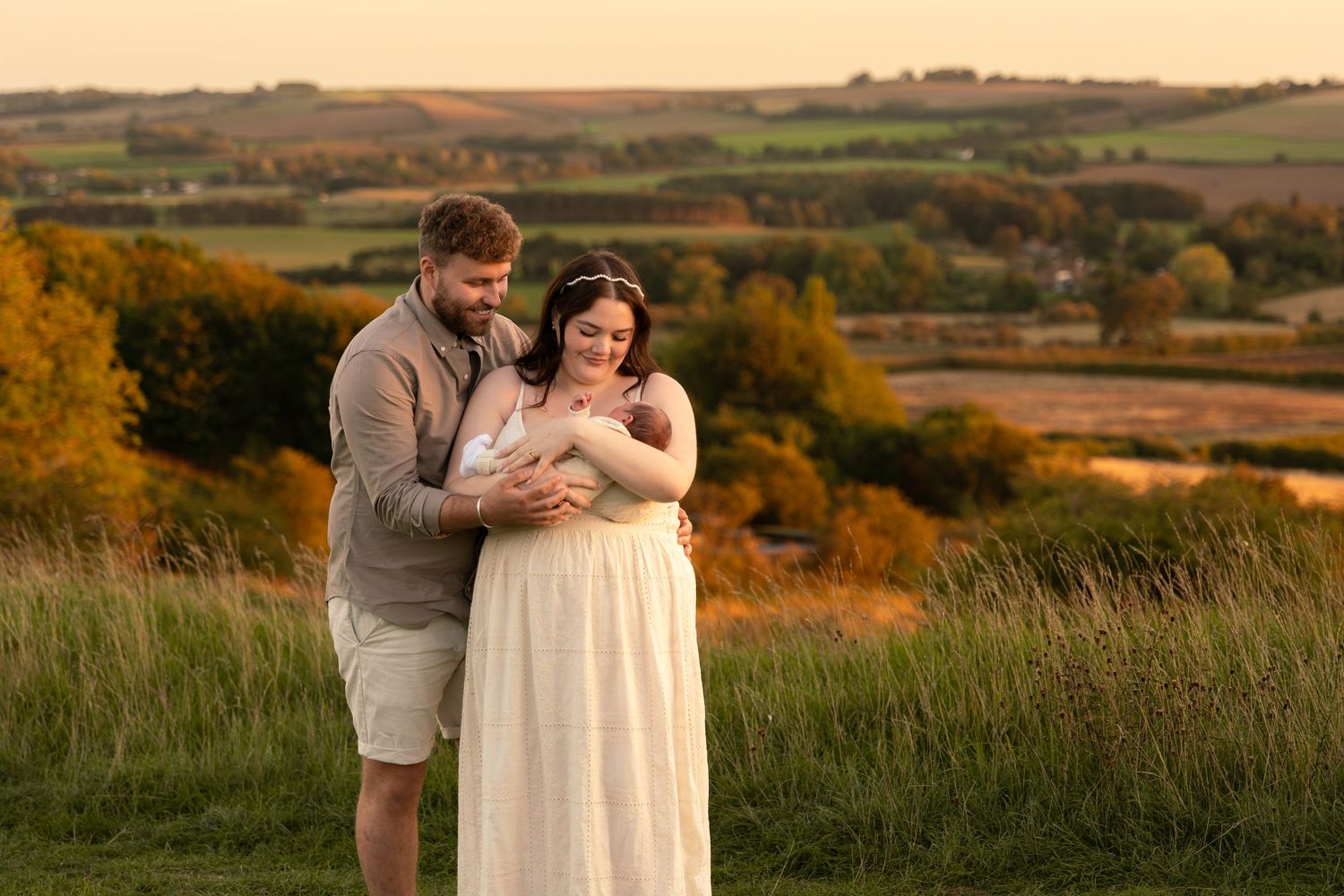 Couple holding newborn baby in golden hour light with rolling countryside hills in background.