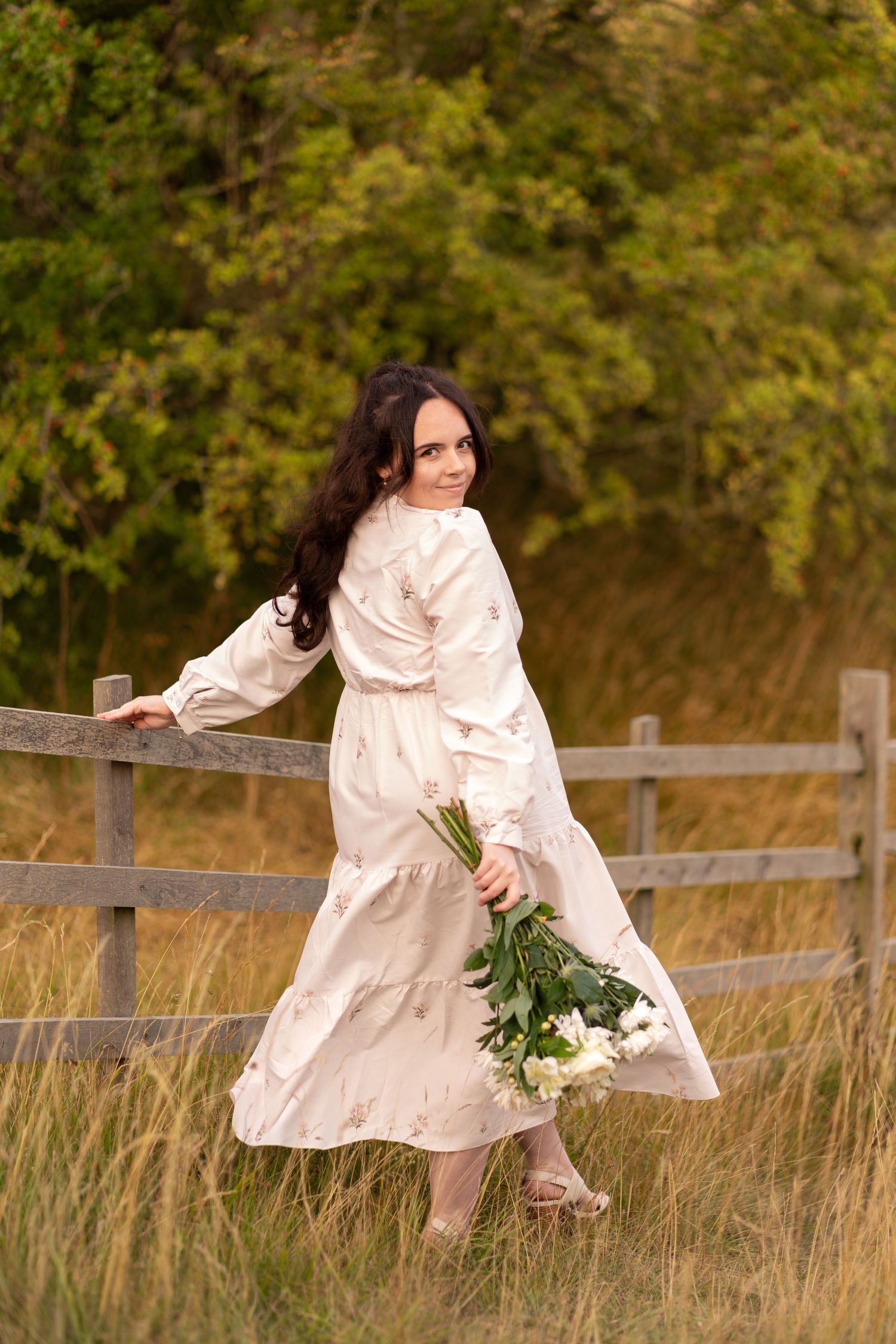 Woman in white dress holding white flowers by a wooden fence in a golden field with green trees.