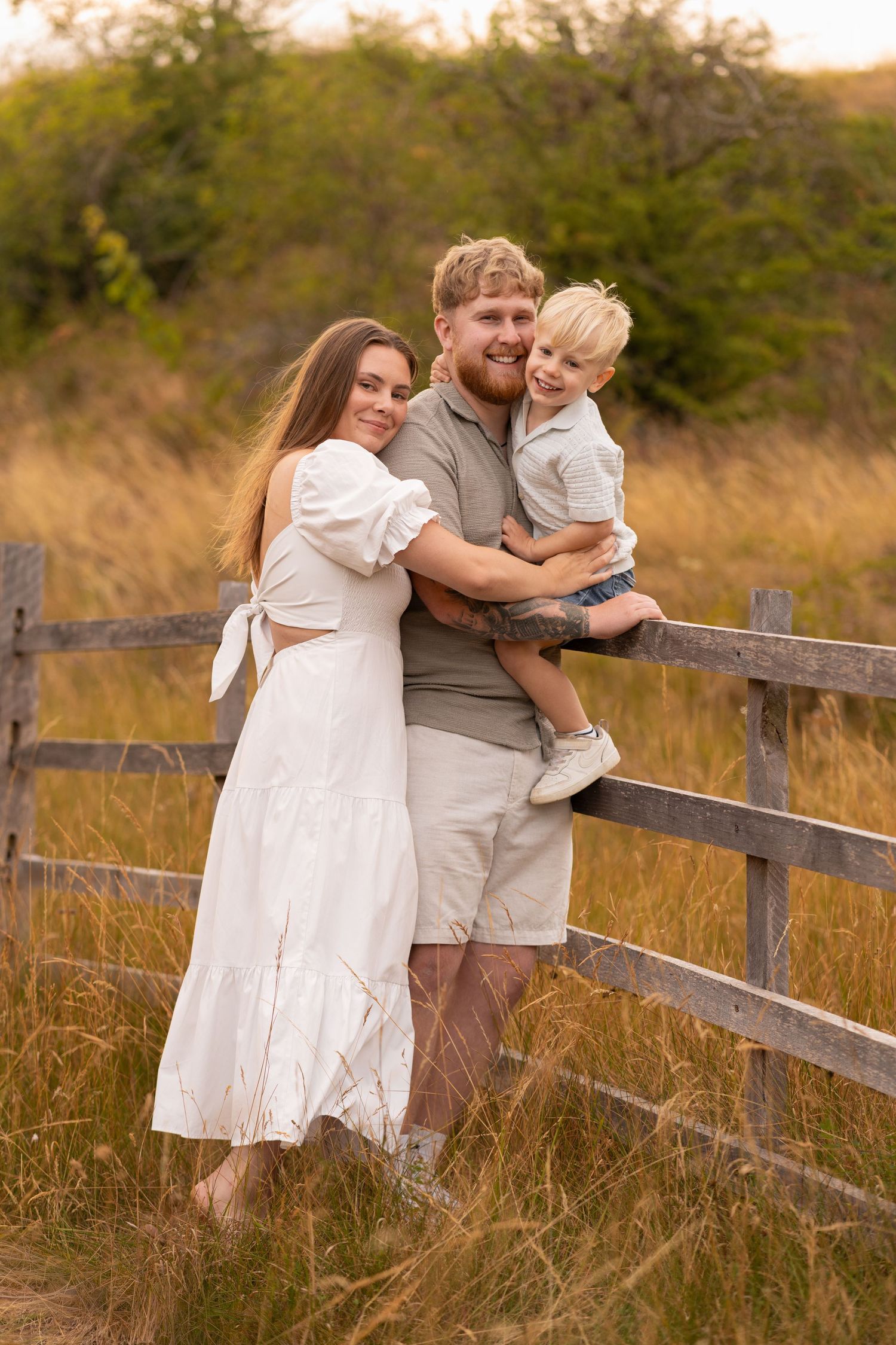 Happy family of three posing by a rustic wooden fence in a golden field, parents smiling with young toddler.