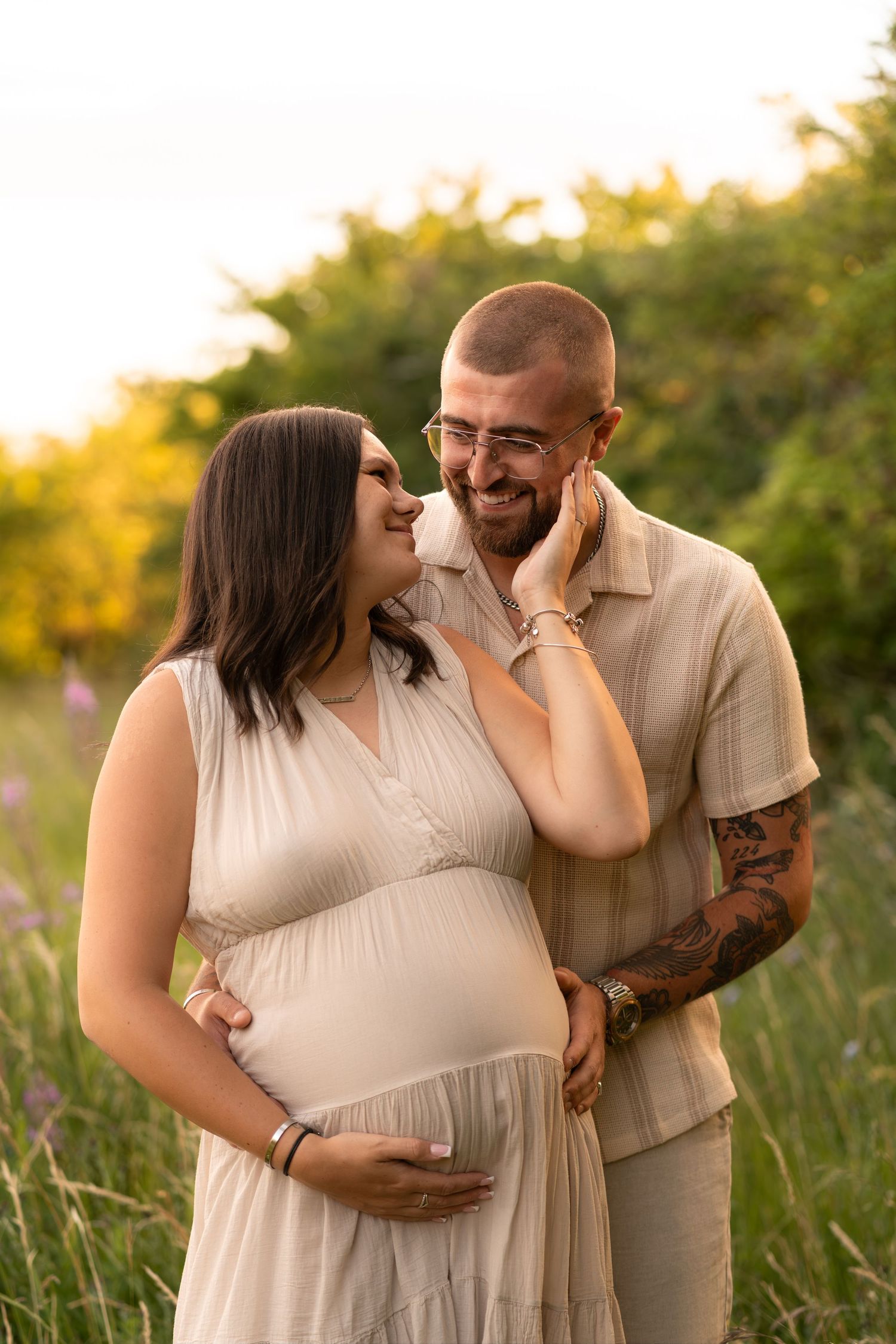 Pregnant couple embracing outdoors at golden hour, woman in beige dress cradling belly, tattooed man in matching outfit smiling warmly.