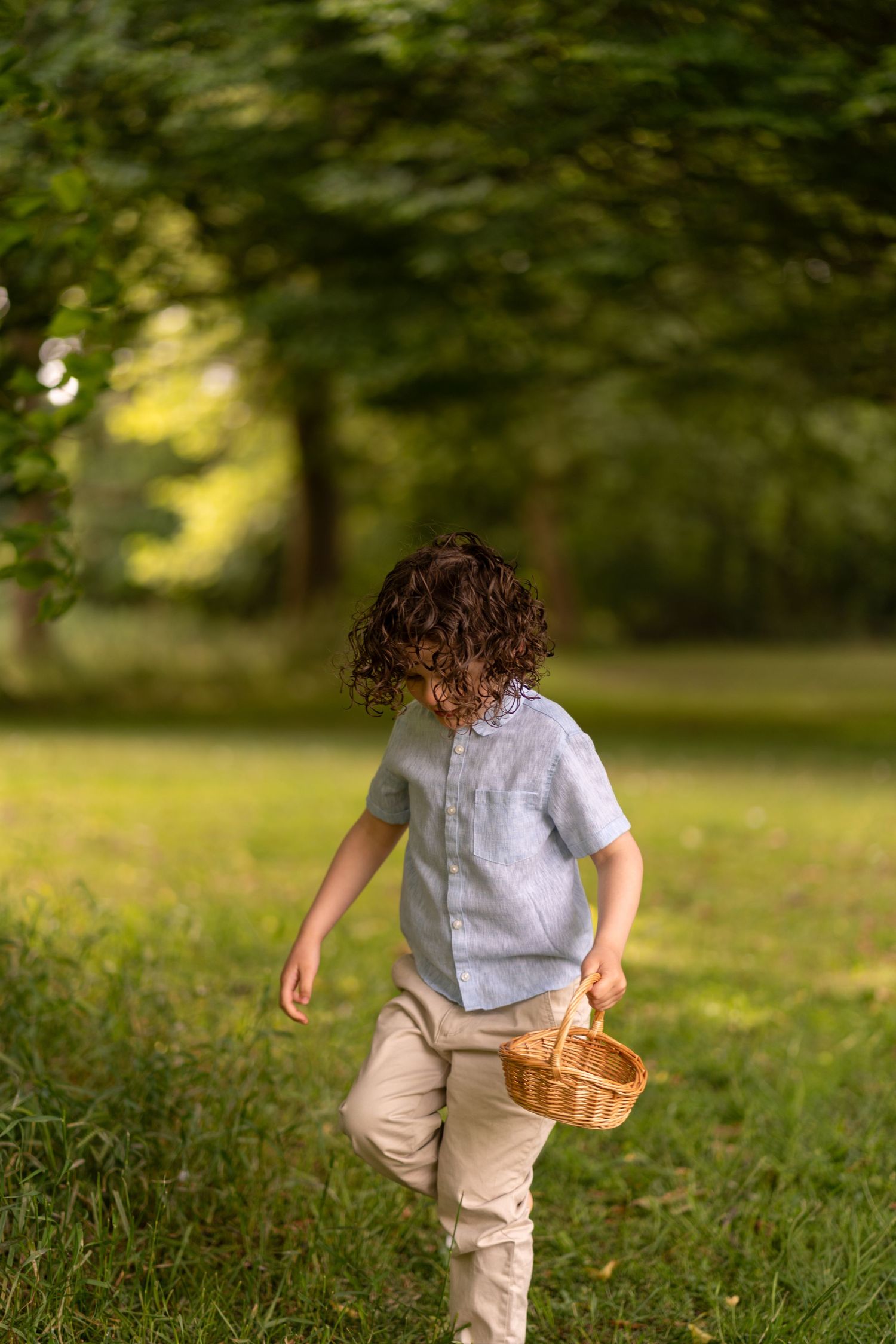 Curly-haired child carrying a wicker basket while walking through a lush green park on a sunny day.