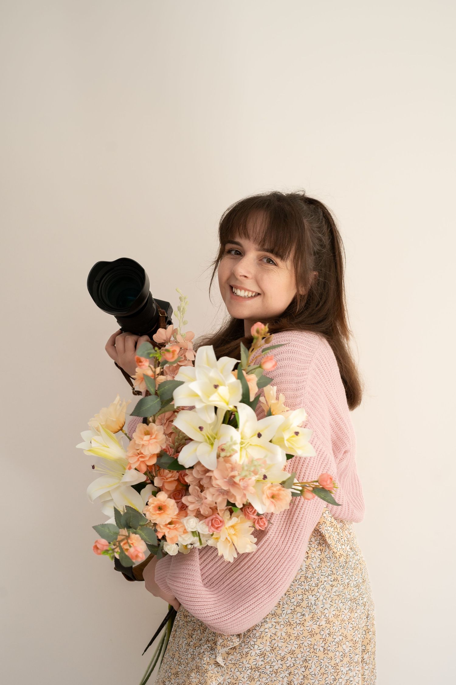 Smiling young woman holding a camera and large bouquet of white and peach flowers, wearing a pink sweater and floral skirt.