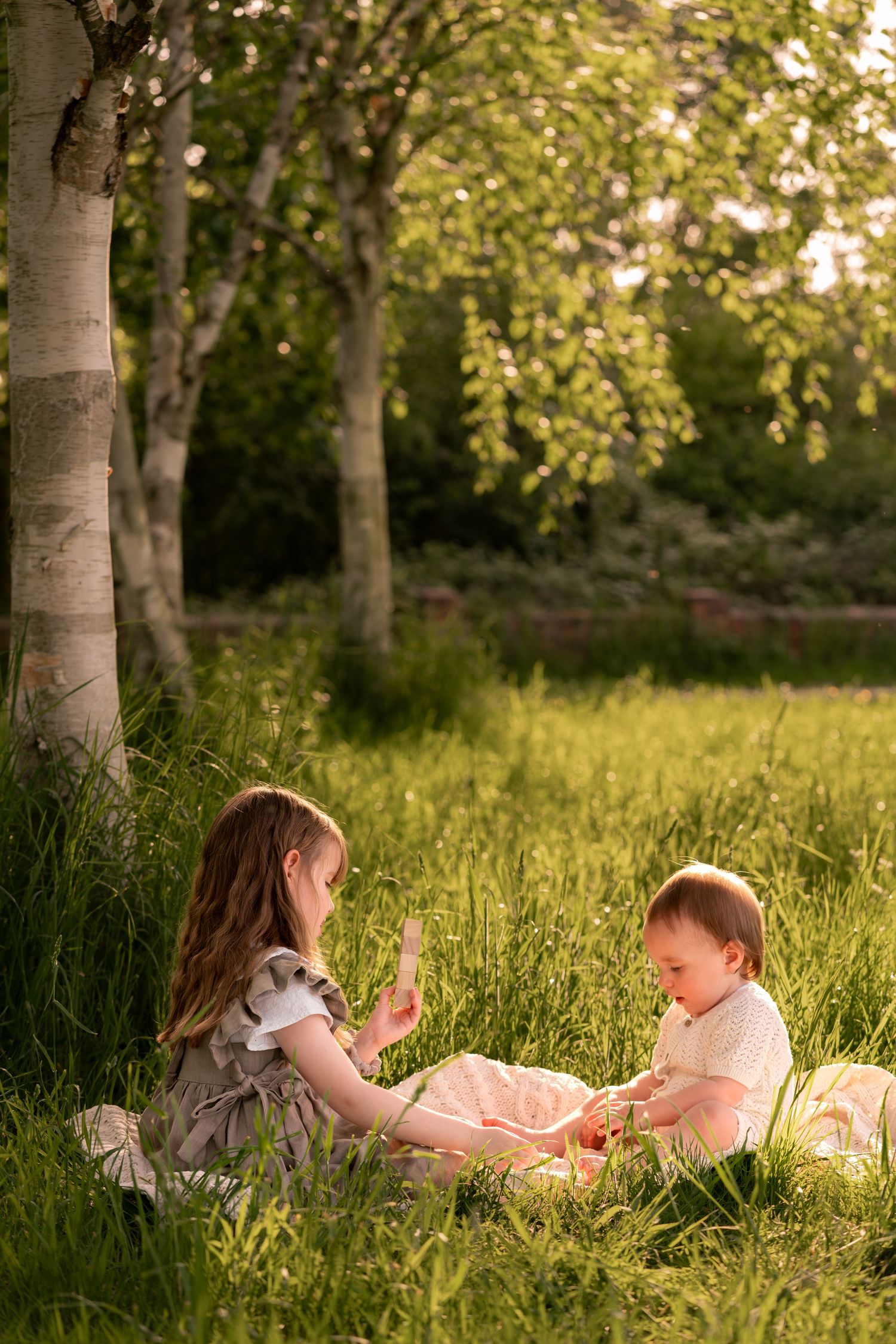 Two young children sitting in lush green grass near birch trees during golden hour sunset light.