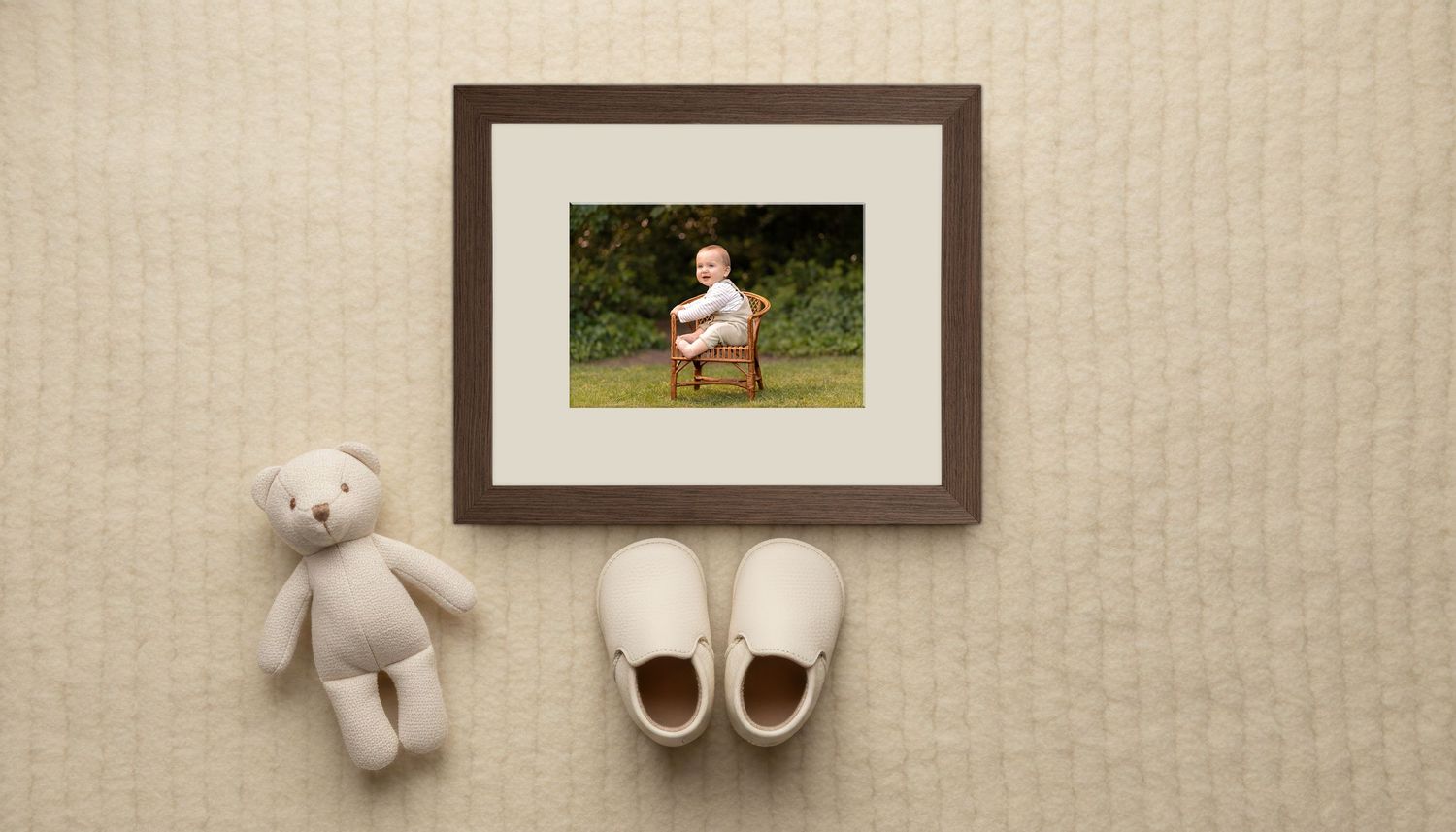 Framed baby photo with a stuffed teddy bear and white baby shoes on a cream textured background.
