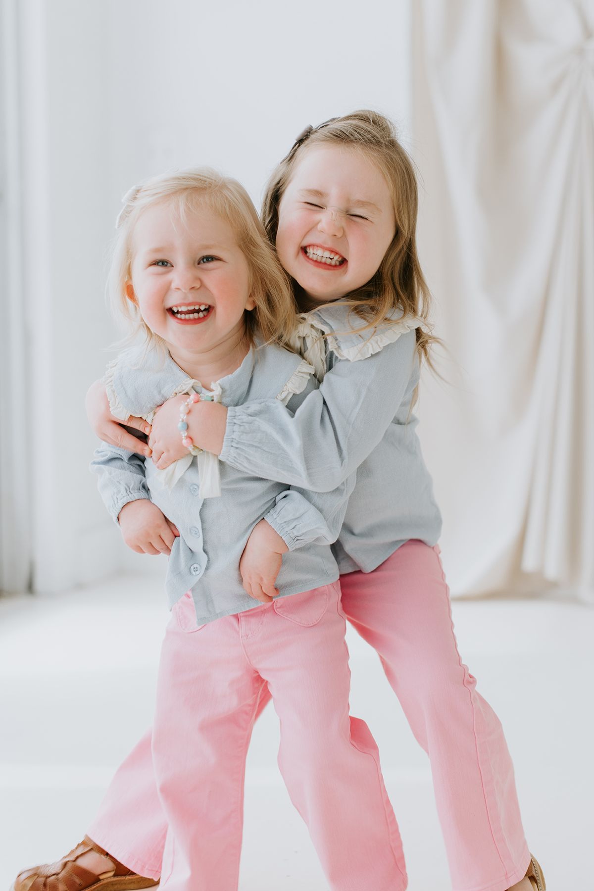 Two children wearing matching chambray tops and pink pants laugh and hug while posing in front of a white backdrop.
