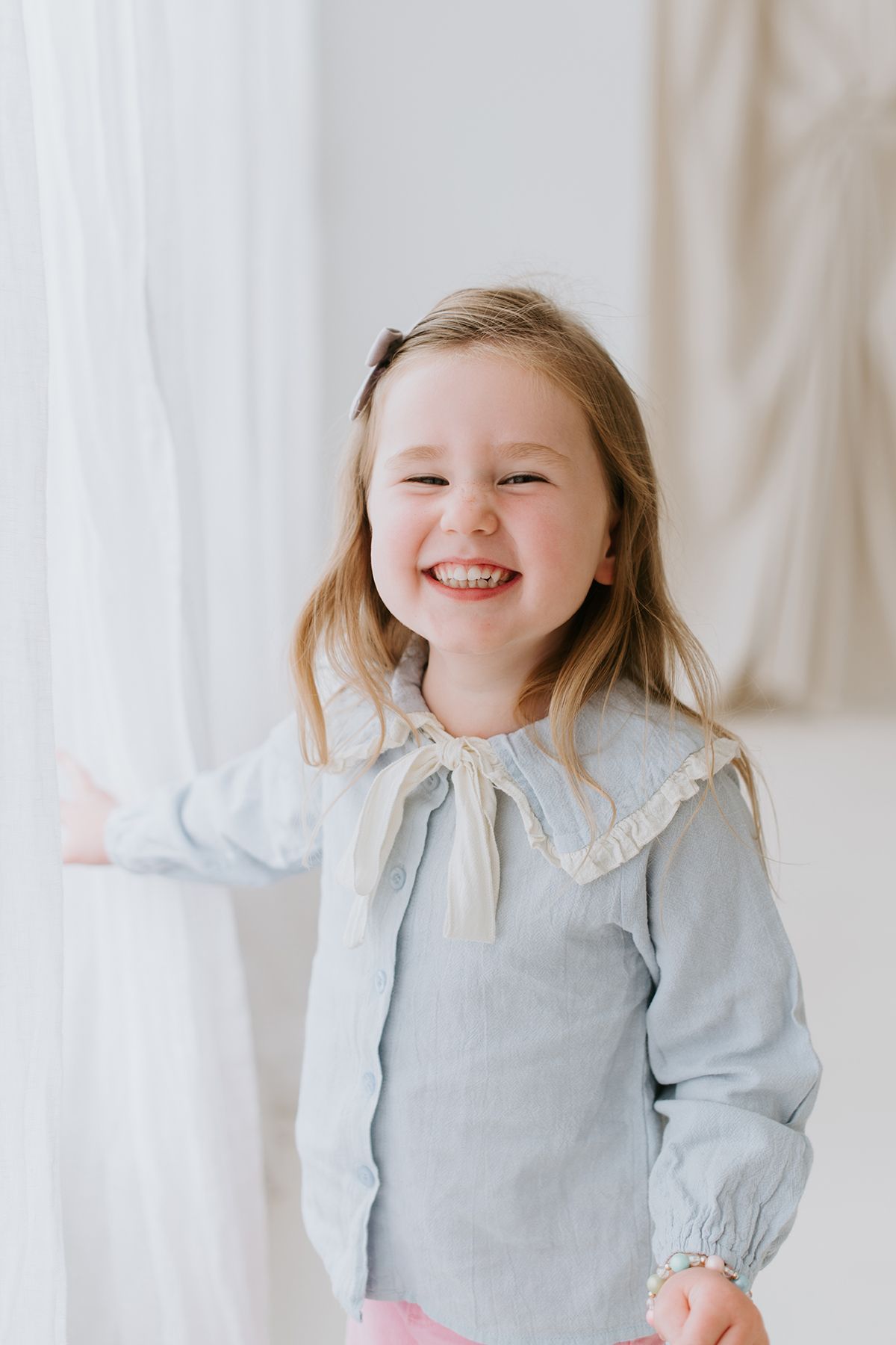 A child wearing a light blue dress with a Peter Pan collar smiles joyfully in front of white curtains.