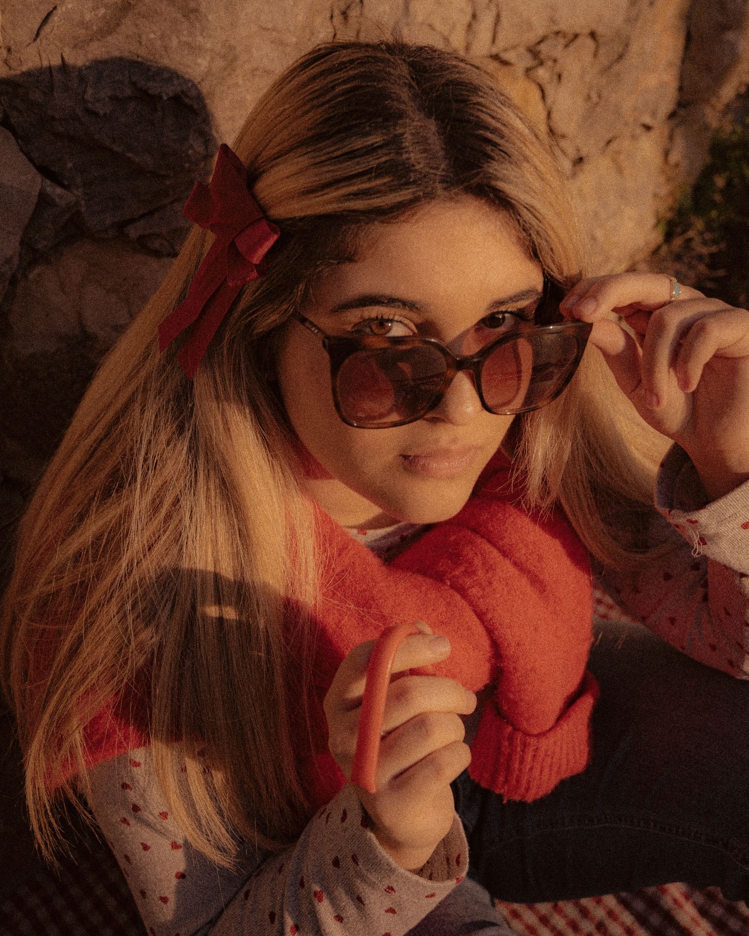 Portrait of someone in sunglasses and red sweater posed against a brown wall in moody lighting.