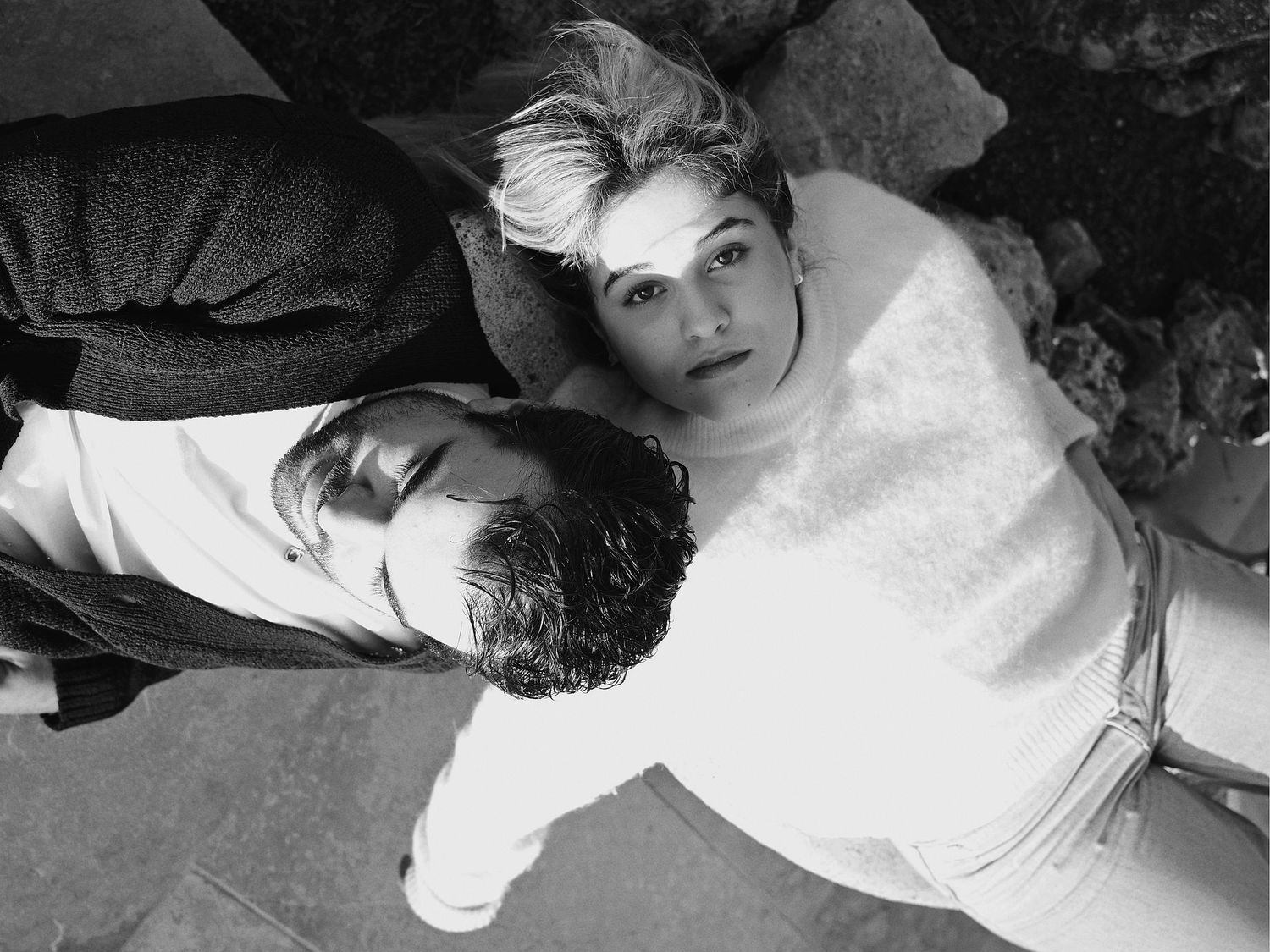 A black and white artistic portrait of two people lying down and looking up at the camera.