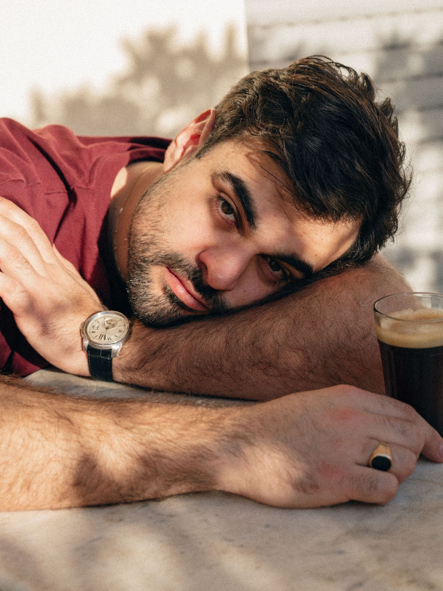 A tired person in a burgundy shirt rests their head on a counter while wearing a wristwatch.