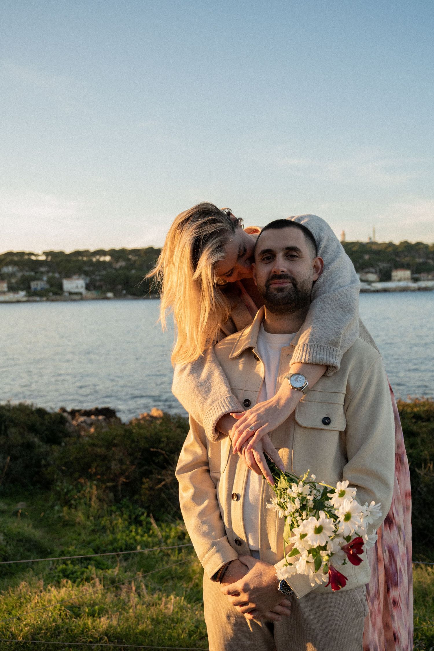 A couple embraces by the waterfront at sunset, with the woman holding a bouquet of white and red flowers.