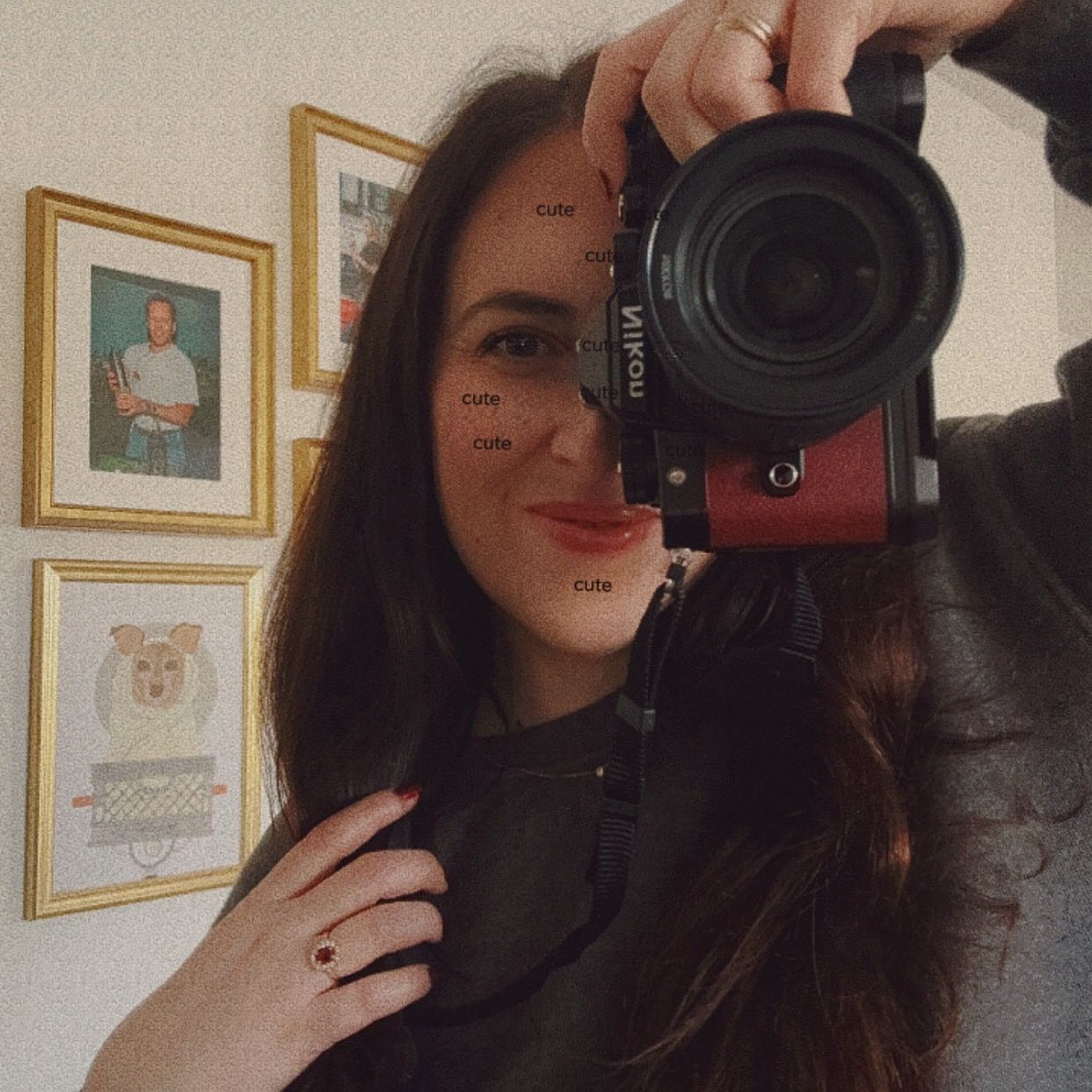 A photographer with long dark hair takes a selfie with a professional camera in front of framed artwork on a wall.