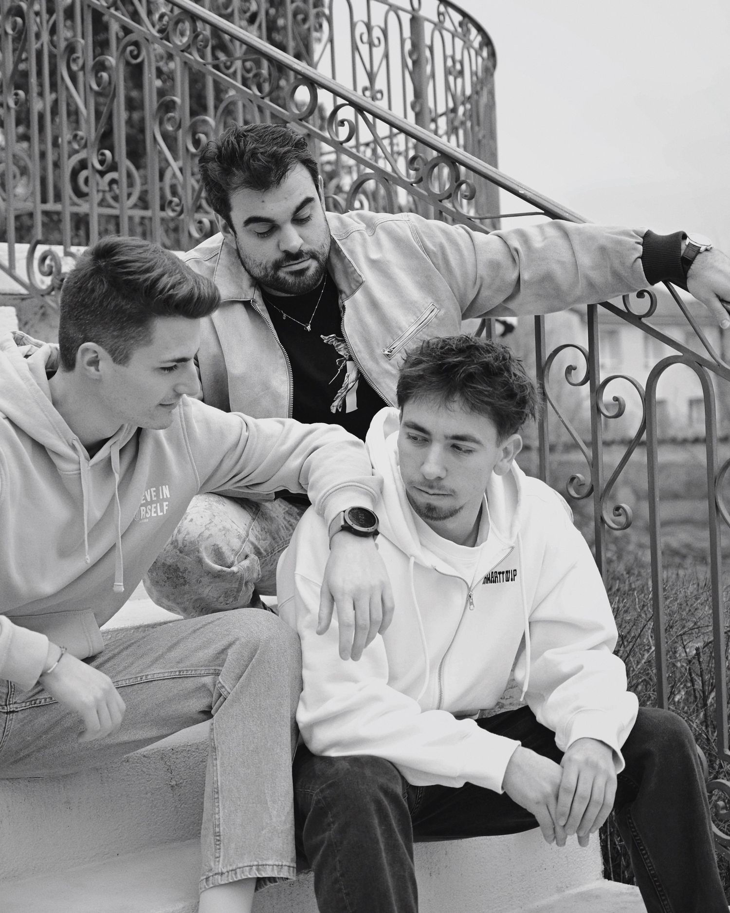 Three young men in casual attire lounge together on ornate iron stairs in this vintage black and white photograph.