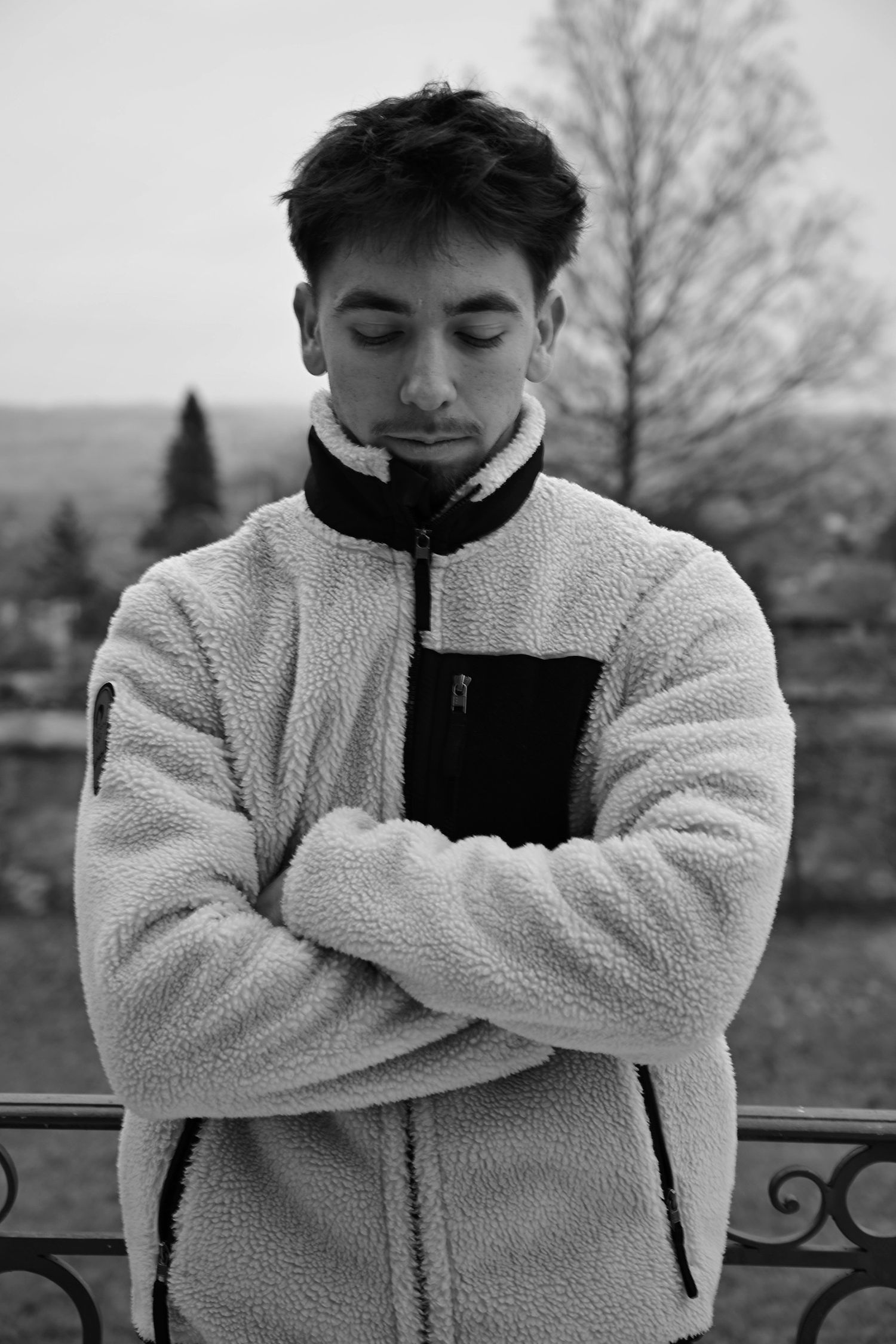 Black and white portrait of someone in a cozy fleece jacket with arms crossed standing outdoors against a scenic backdrop.
