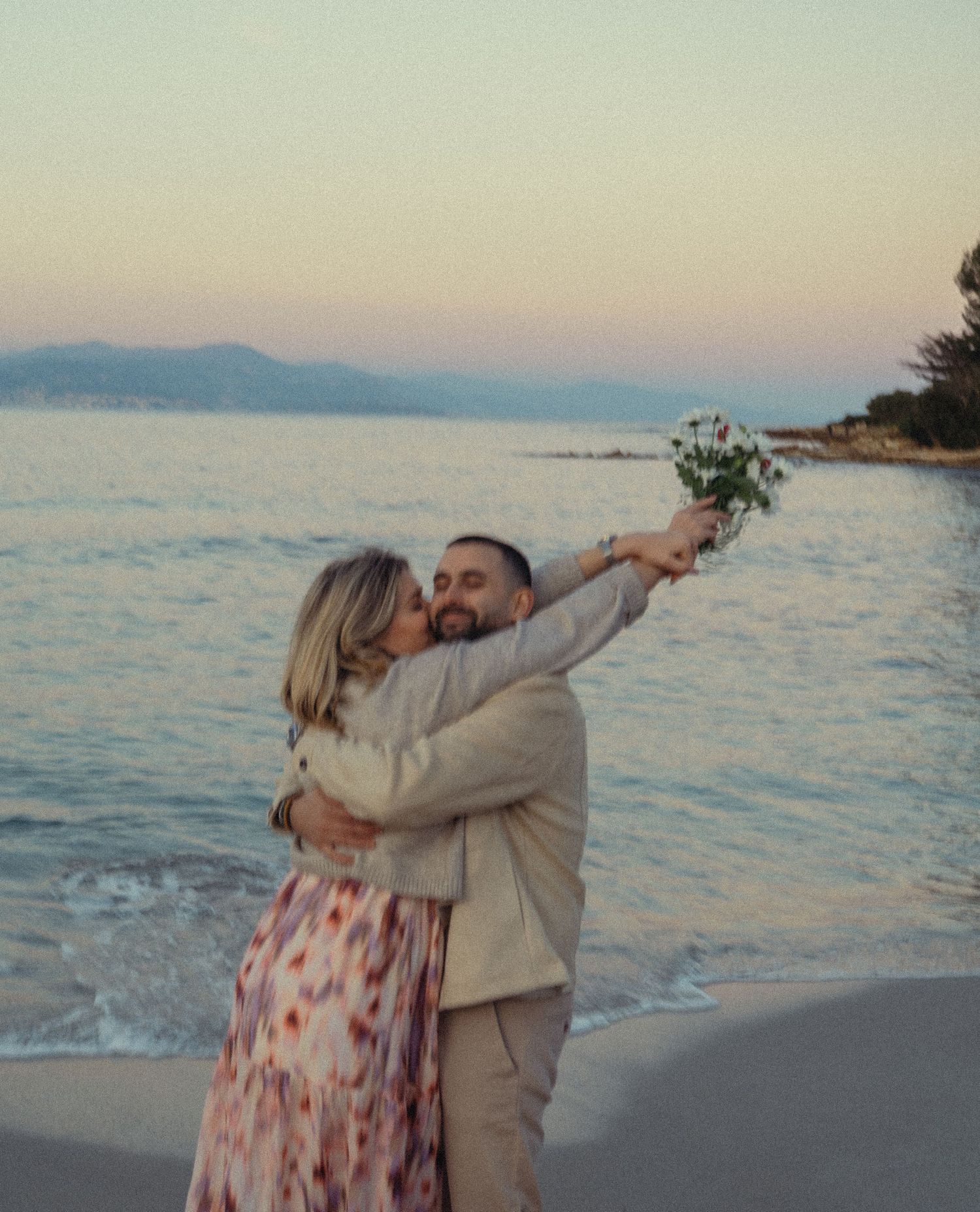 A couple embraces on a beach at sunset with mountains and ocean in the background.
