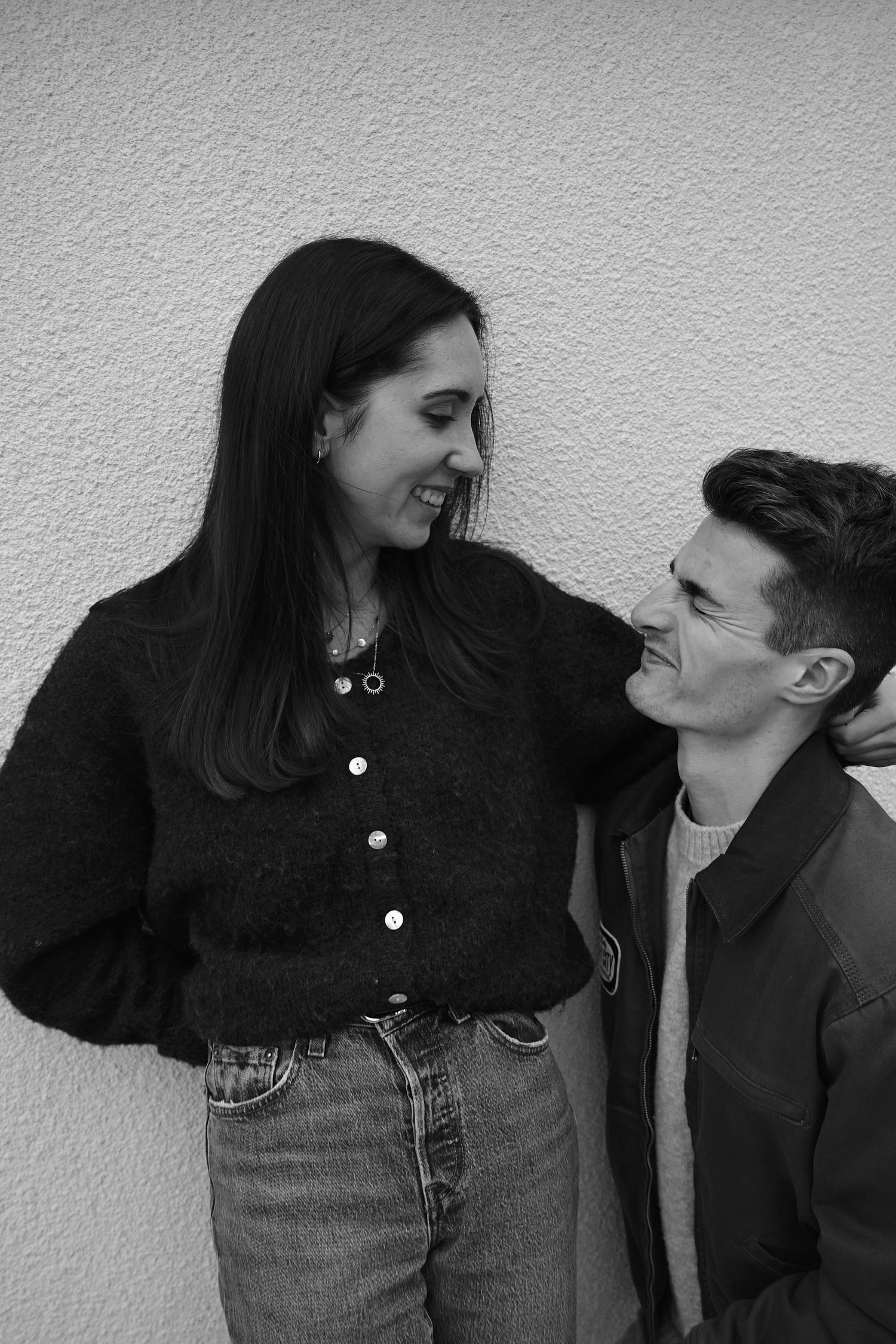 A casual couple sharing a tender moment against a white wall in black and white.