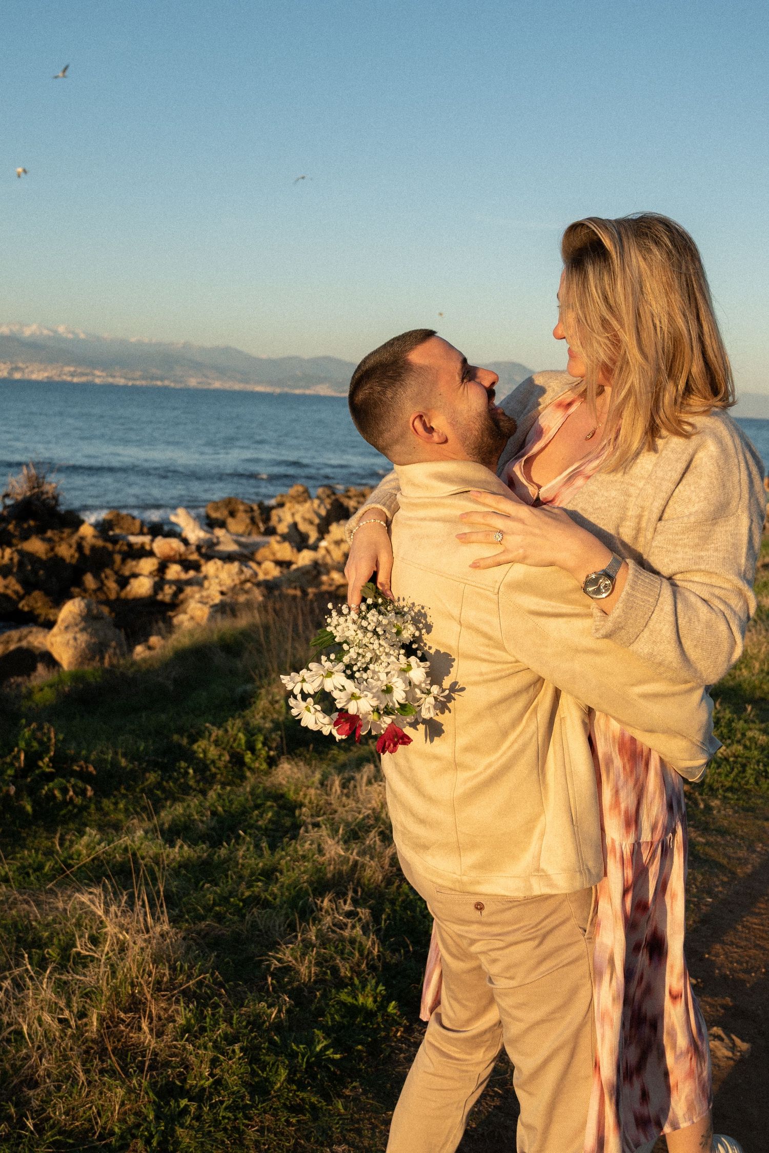 A romantic couple embracing by the rocky coastline at sunset with a bouquet of flowers.