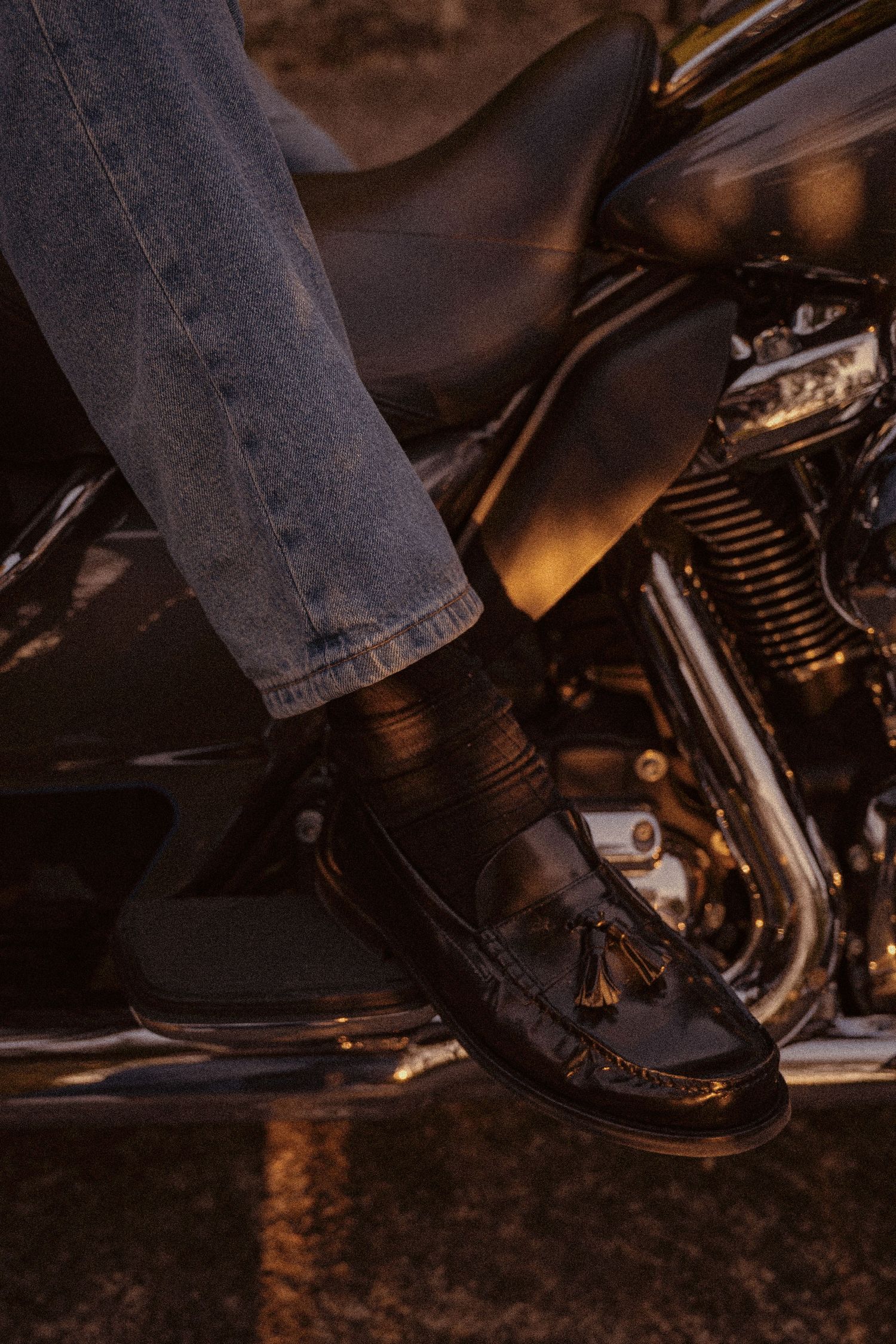 Close-up of black leather loafer on motorcycle footrest with denim jeans and chrome details visible.