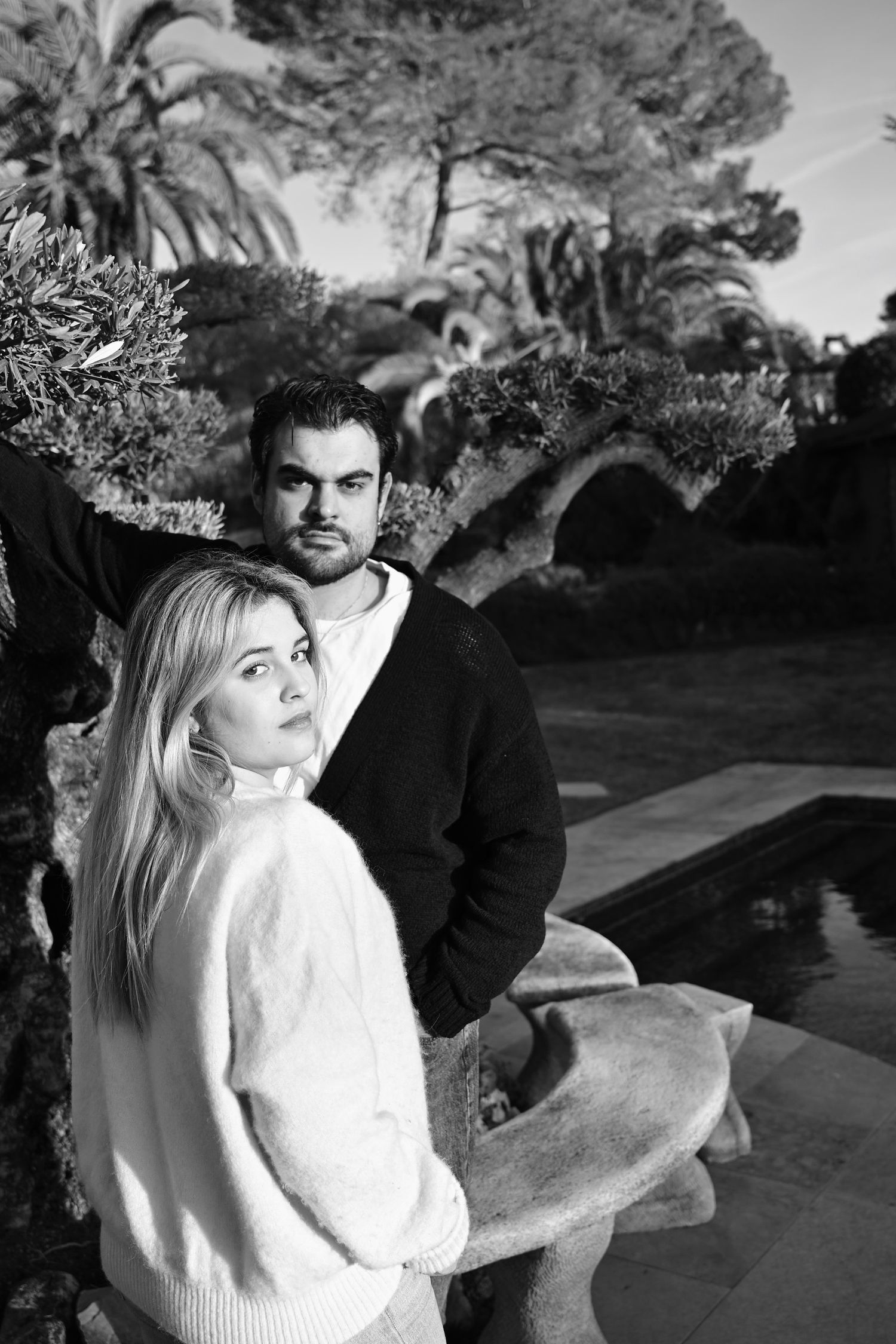 A couple in black and white portrait pose outside near a pool, with palm trees and lush greenery in the background.