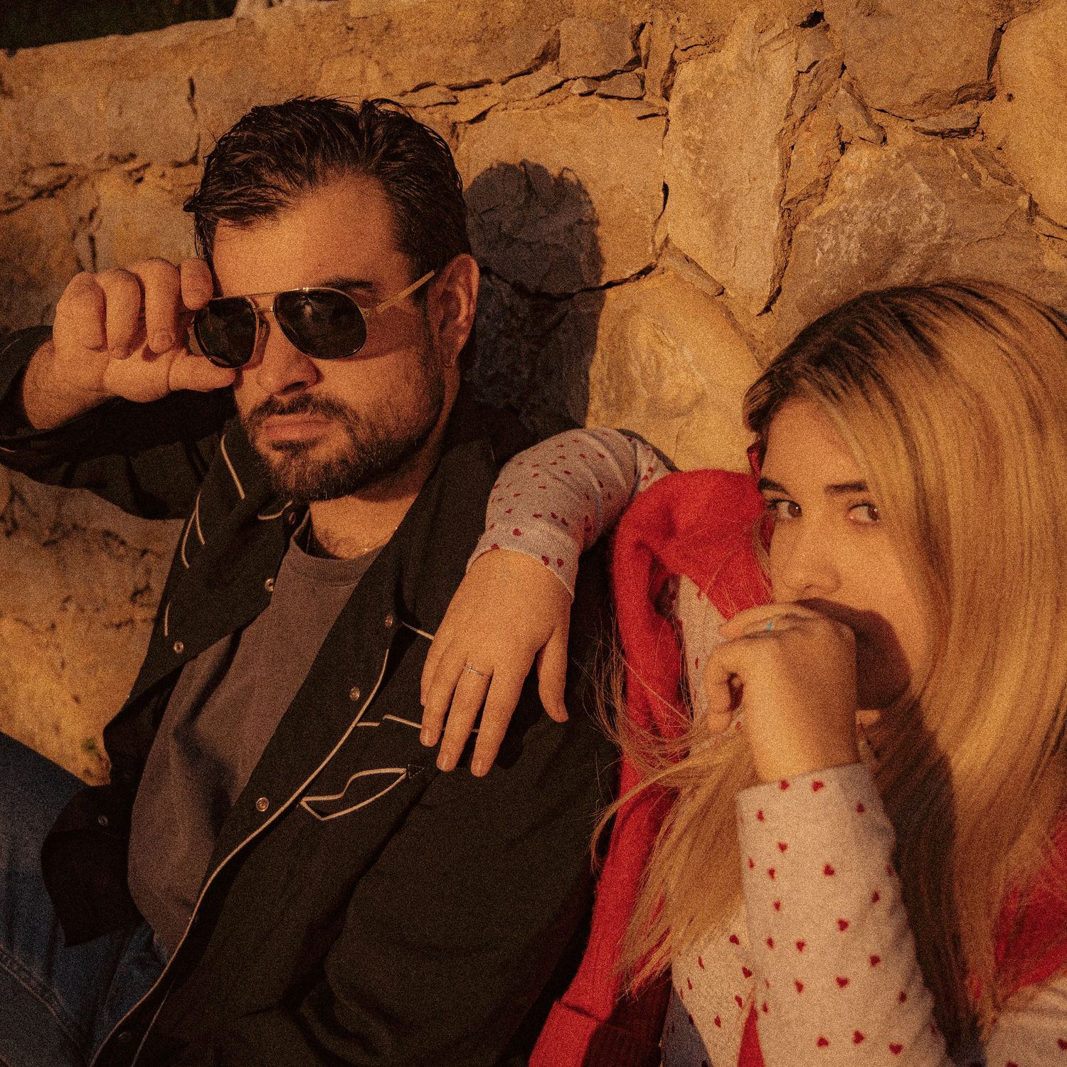 Two people in casual attire pose against a stone wall at sunset, with one wearing sunglasses and dark clothing.