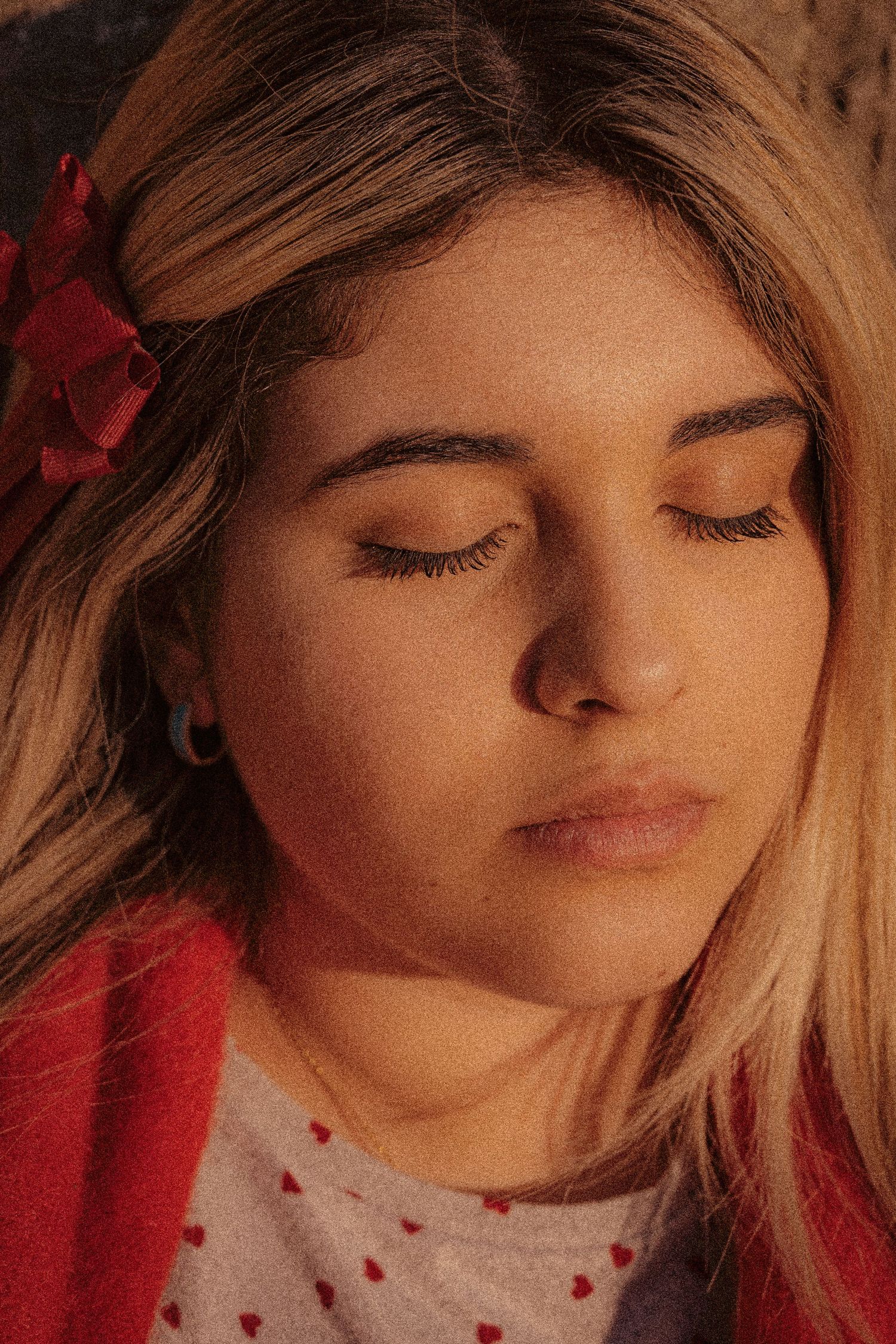 A woman with closed eyes in warm lighting wears a white and red polka dot top with a red flower in her blonde hair.