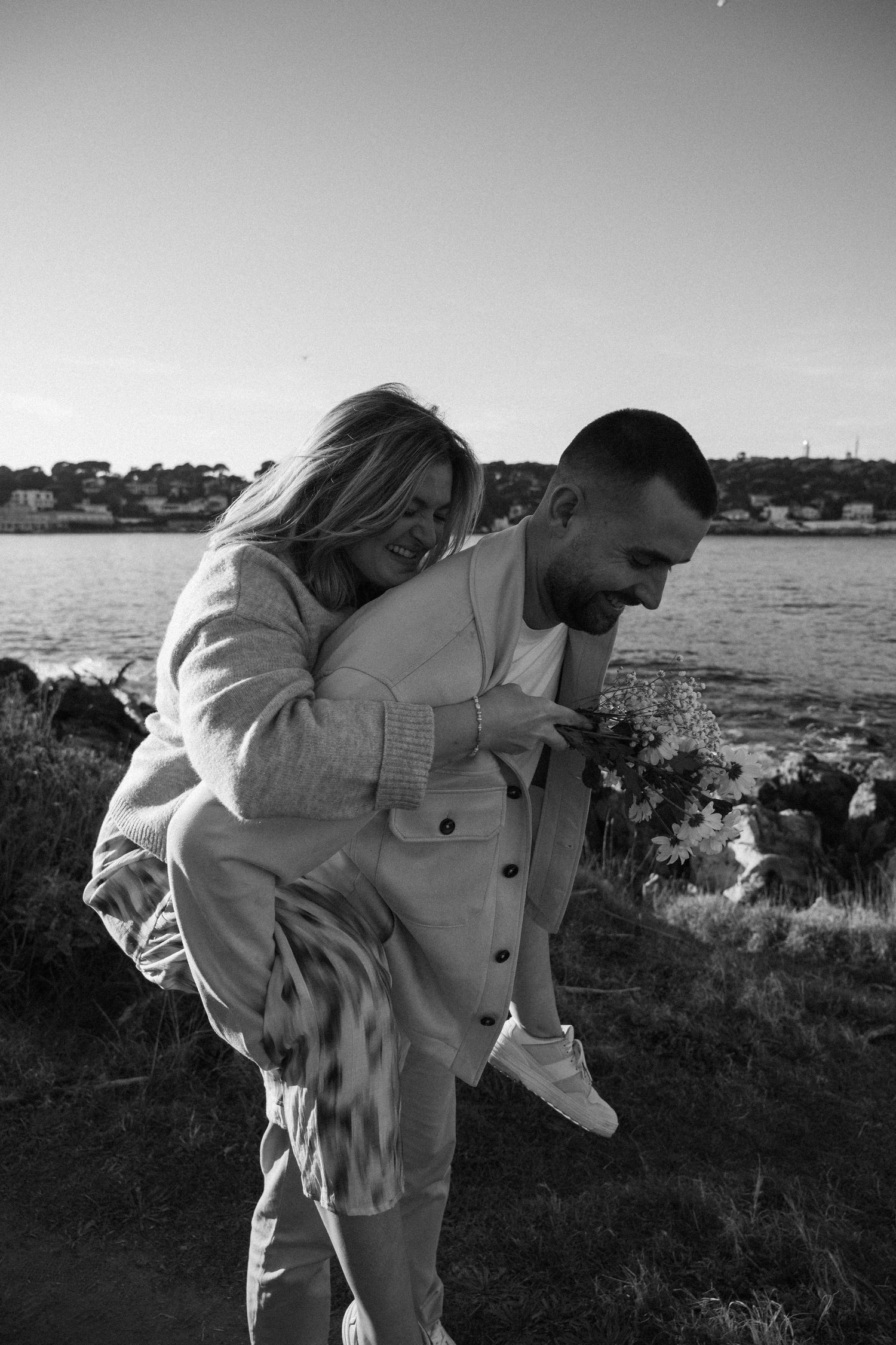 A couple shares a joyful piggyback moment near the waterfront in black and white photography.