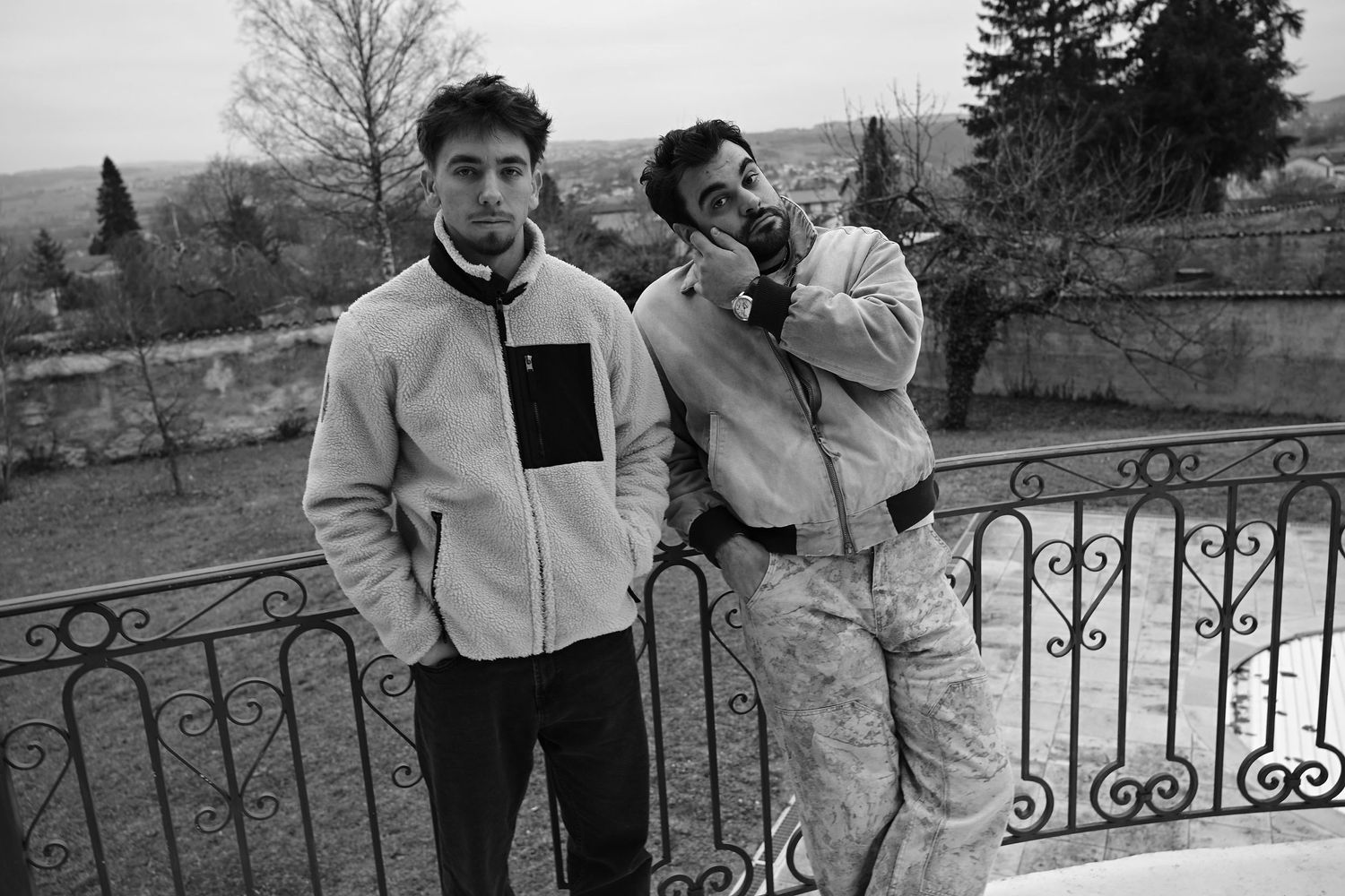 Two individuals in casual outerwear stand on a balcony with wrought iron railing overlooking a scenic winter landscape.