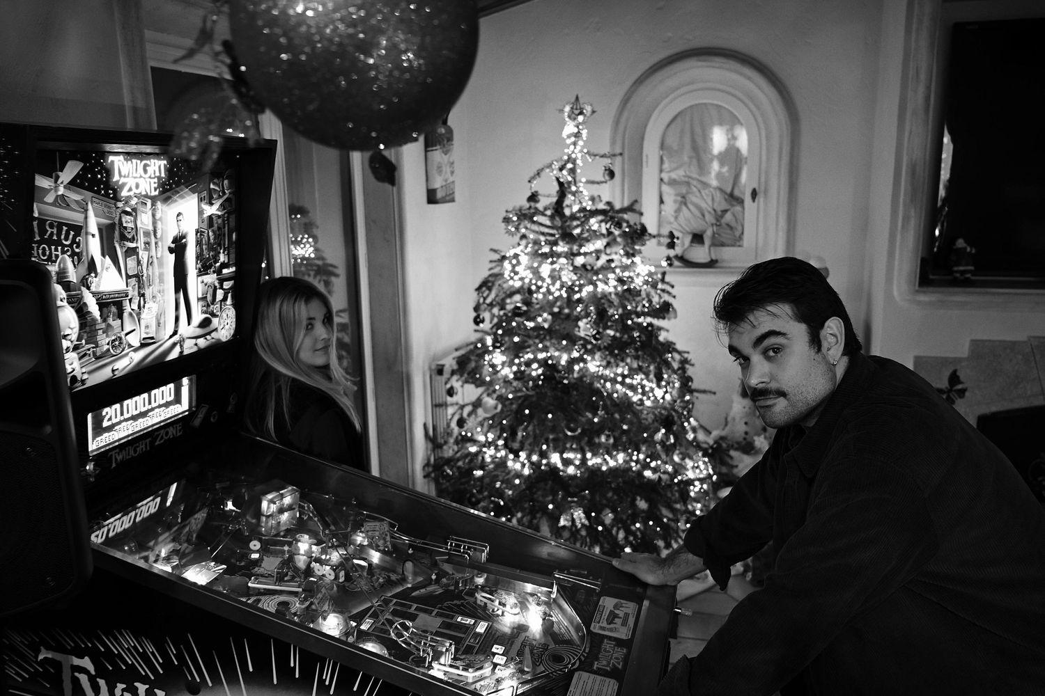 Two people gathered around a Christmas tree with decorative lights in a dimly lit room.