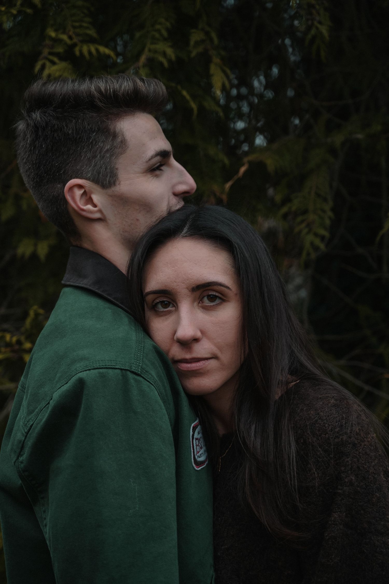 Intimate portrait of couple in outdoor setting with dark moody lighting and green foliage backdrop.