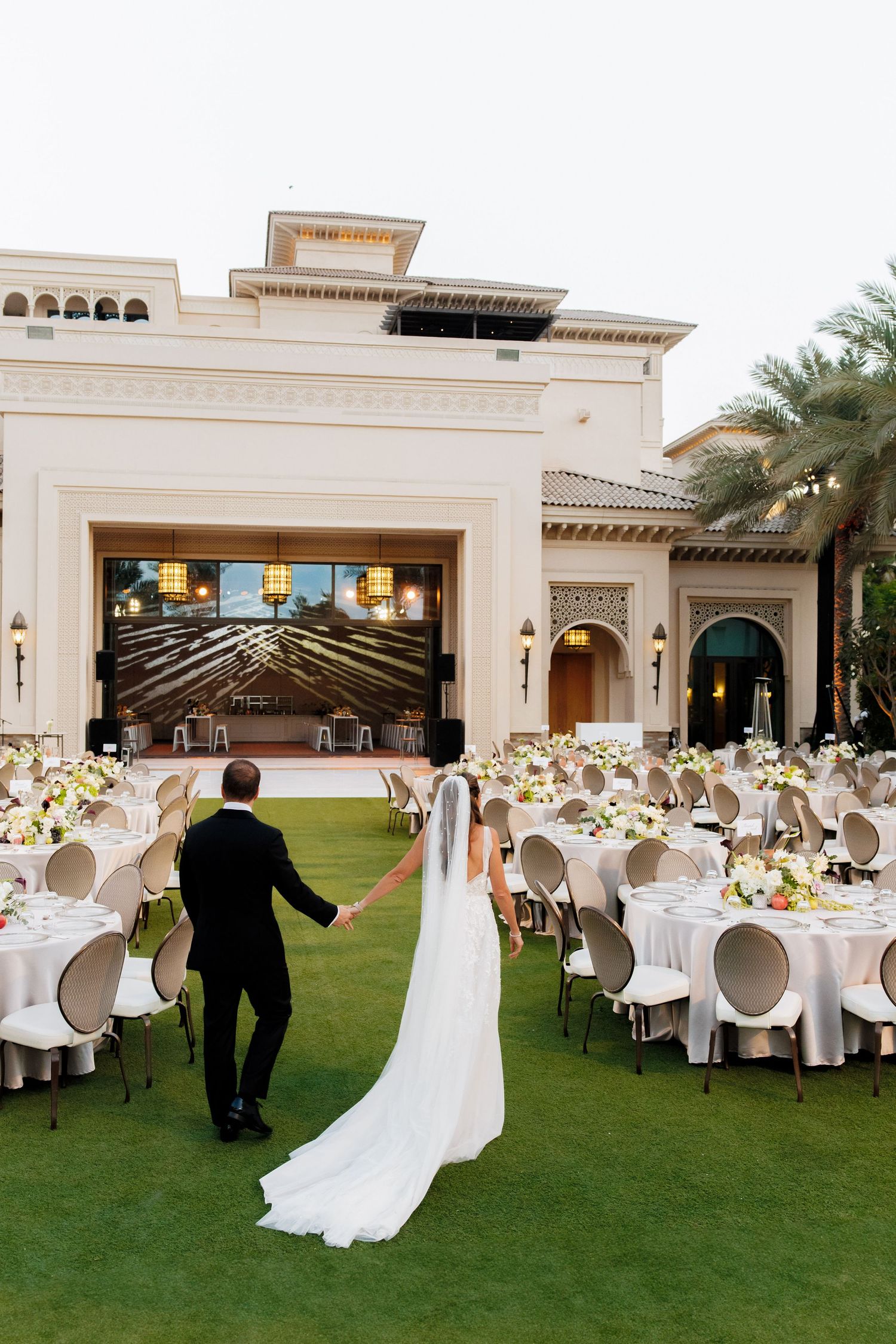 Wedding reception setup with white modern chairs and tables on a manicured lawn by a grand venue.