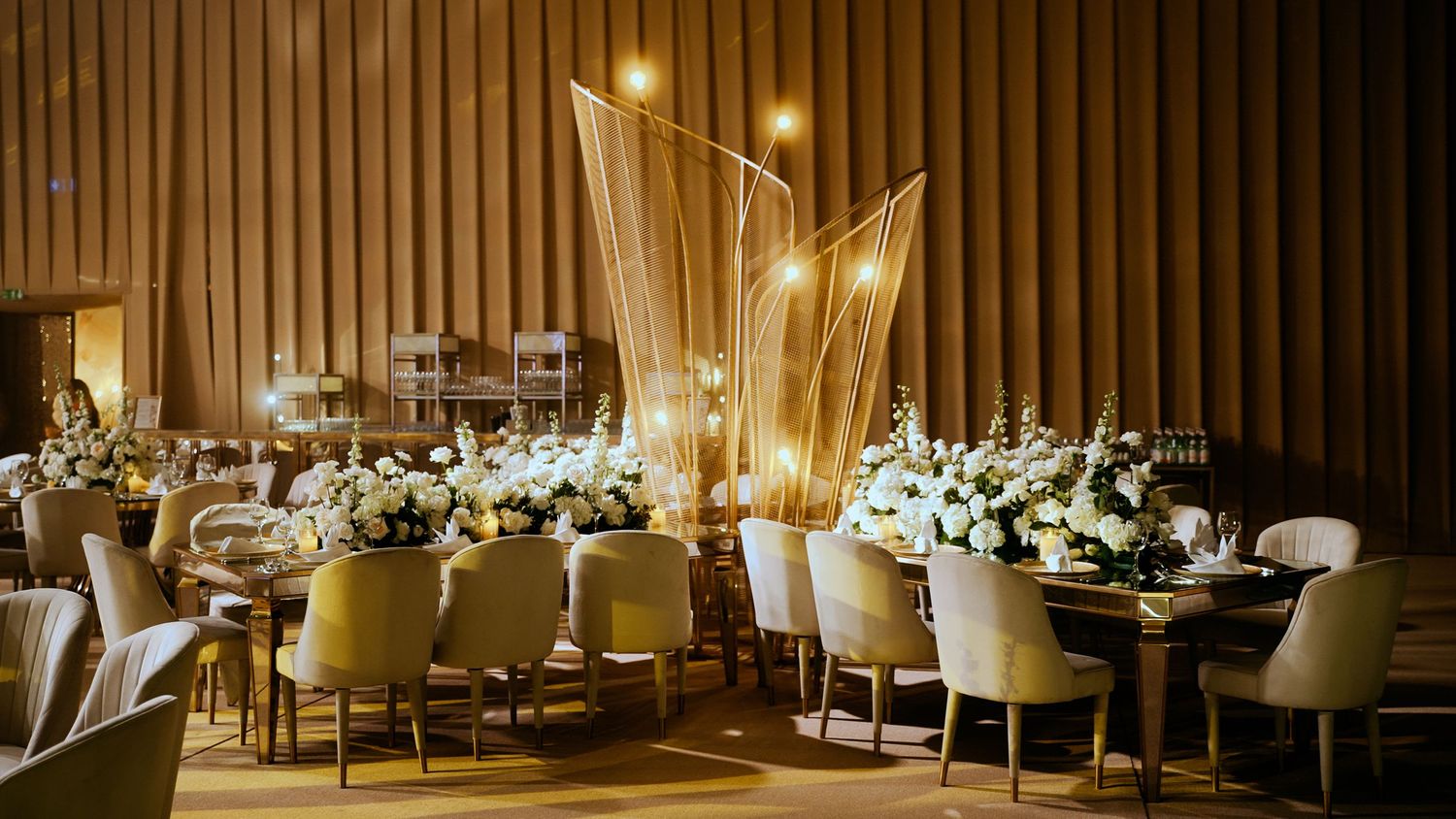 Elegant banquet hall setup with modern gold decor elements and white upholstered chairs in warm ambient lighting.