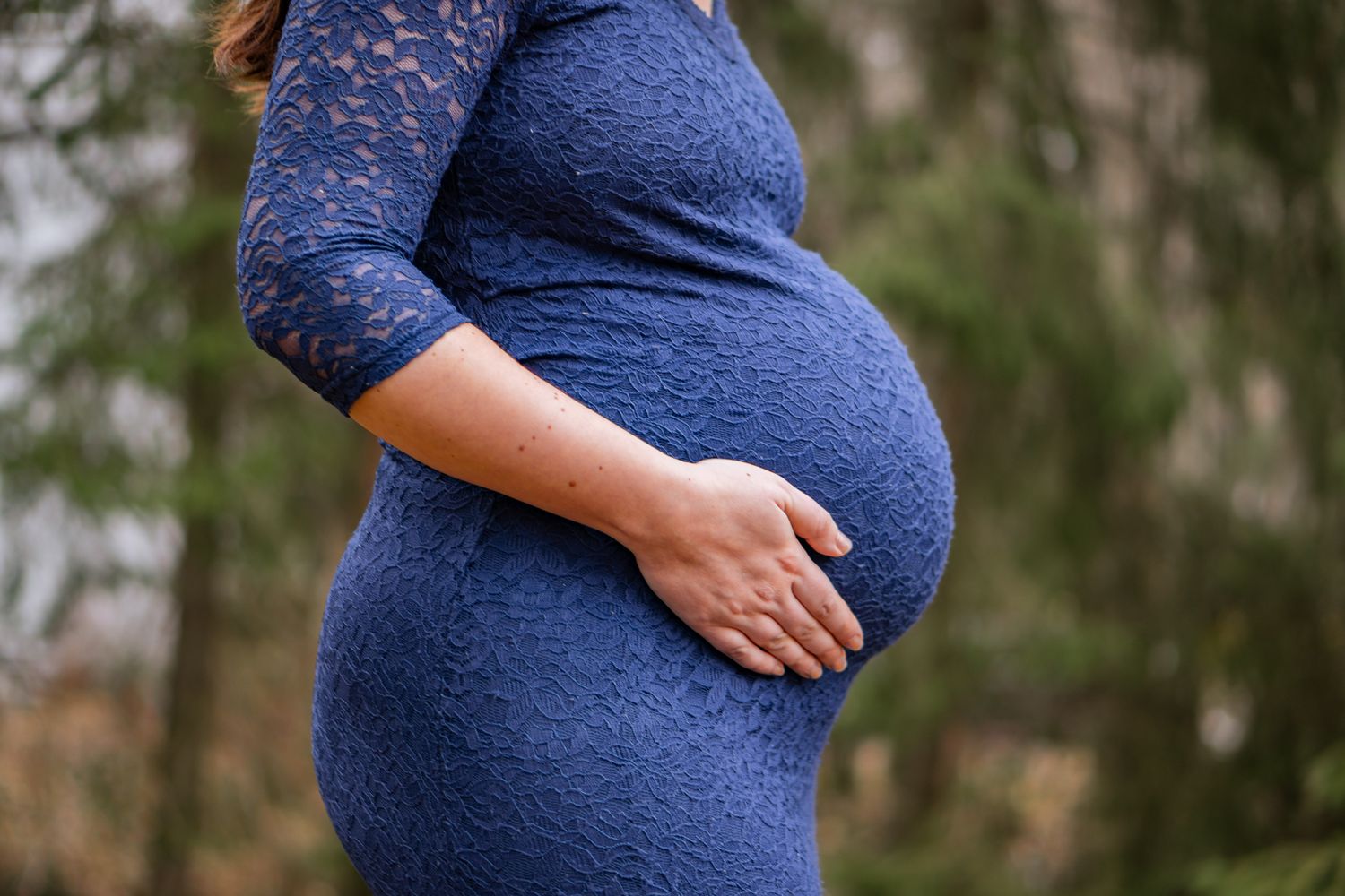 Gravid kvinna i blå spetsklänning håller om sin gravida mage med skogen i bakgrunden. Gravidfotograf, Umeå, Västerbotten