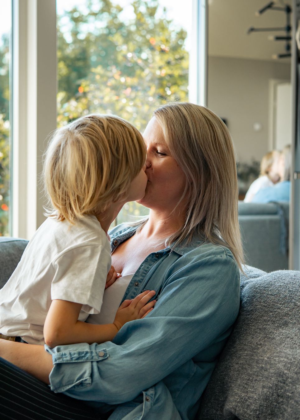 Ett fint ögonblick mellan mamma och son som delar en puss när de sitter i en grå soffa nära soliga fönster. Fotograf, nyföddfotograf, Umeå