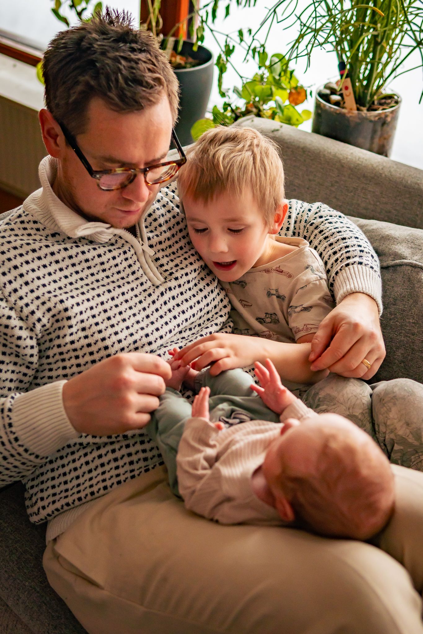 Mysig stund med pappa som sitter i soffan med armen runt sin son och har sin bebis i knät, båda håller i bebis fötter. Fotograf, Umeå