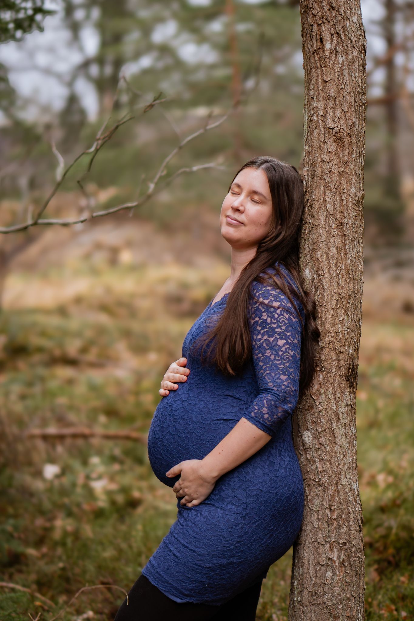 Gravid kvinna i blå spetsklänning lutar sig mot en trädstam i skogen och ler lugnt när hon håller om magen. Gravidfotograf, Umeå