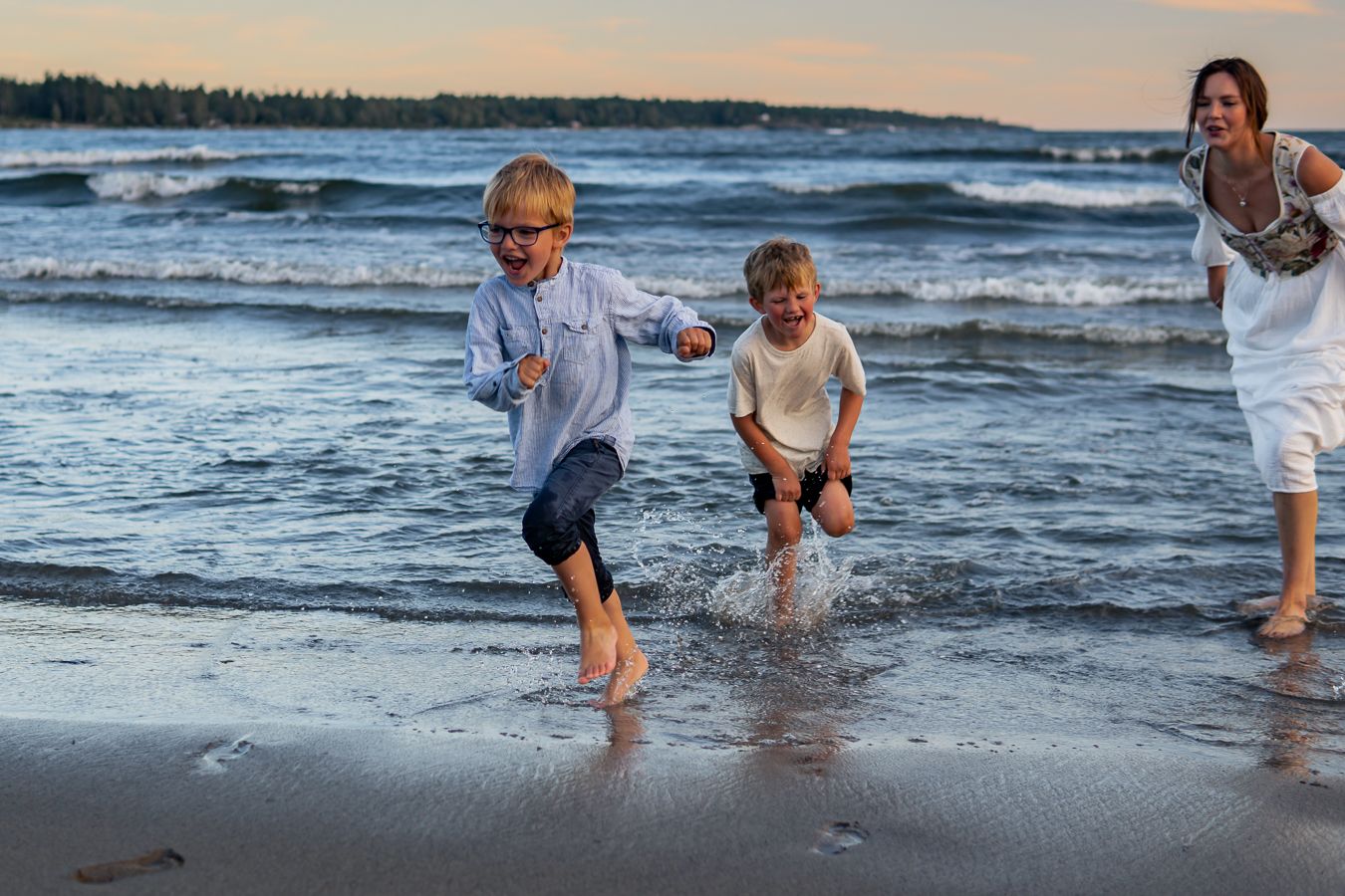 En mamma och två barn springer och leker med kläderna på i vattnat vid strandkanten i solnedgång med havet bakom. Fotograf, Umeå