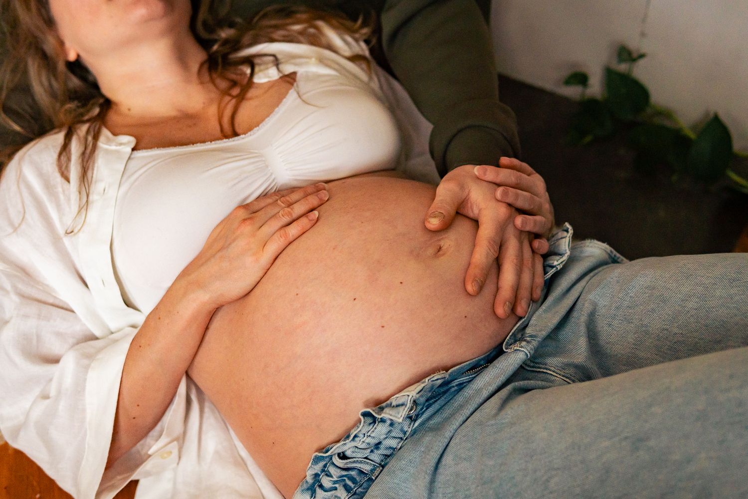 En kvinna i vit uppknäppt skjorta och jeans ligger på rygg med händerna på sin gravida mage i mjukt naturligt ljus. Gravidfotograf, Umeå