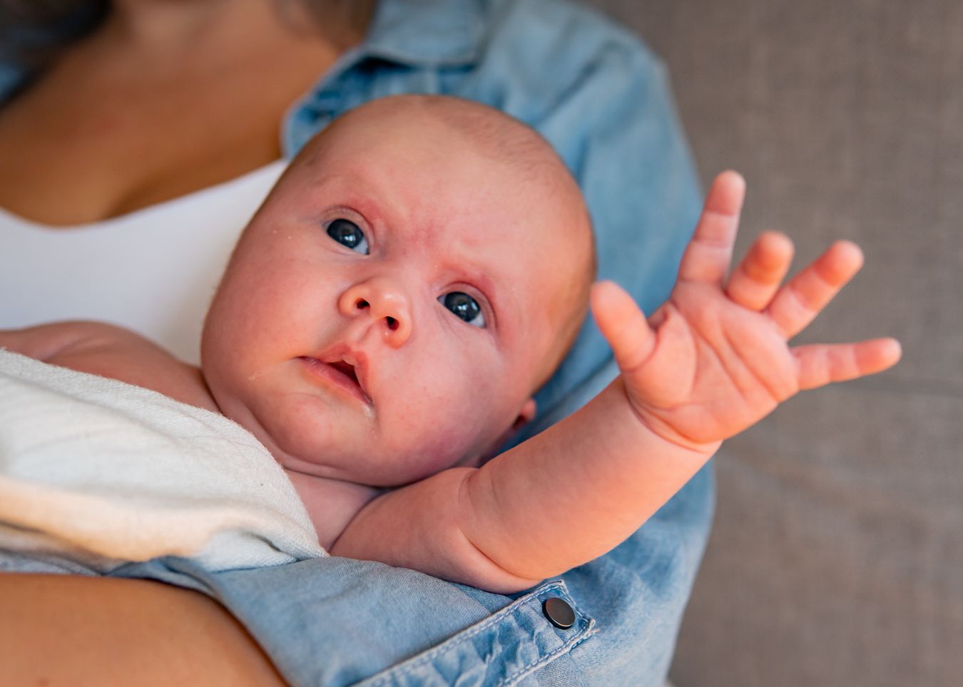 En bebis med blå ögon sträcker ut handen när hon hålls i mammas famn som bär denim. Fotograf, nyföddfotograf, Umeå, Västerbotten