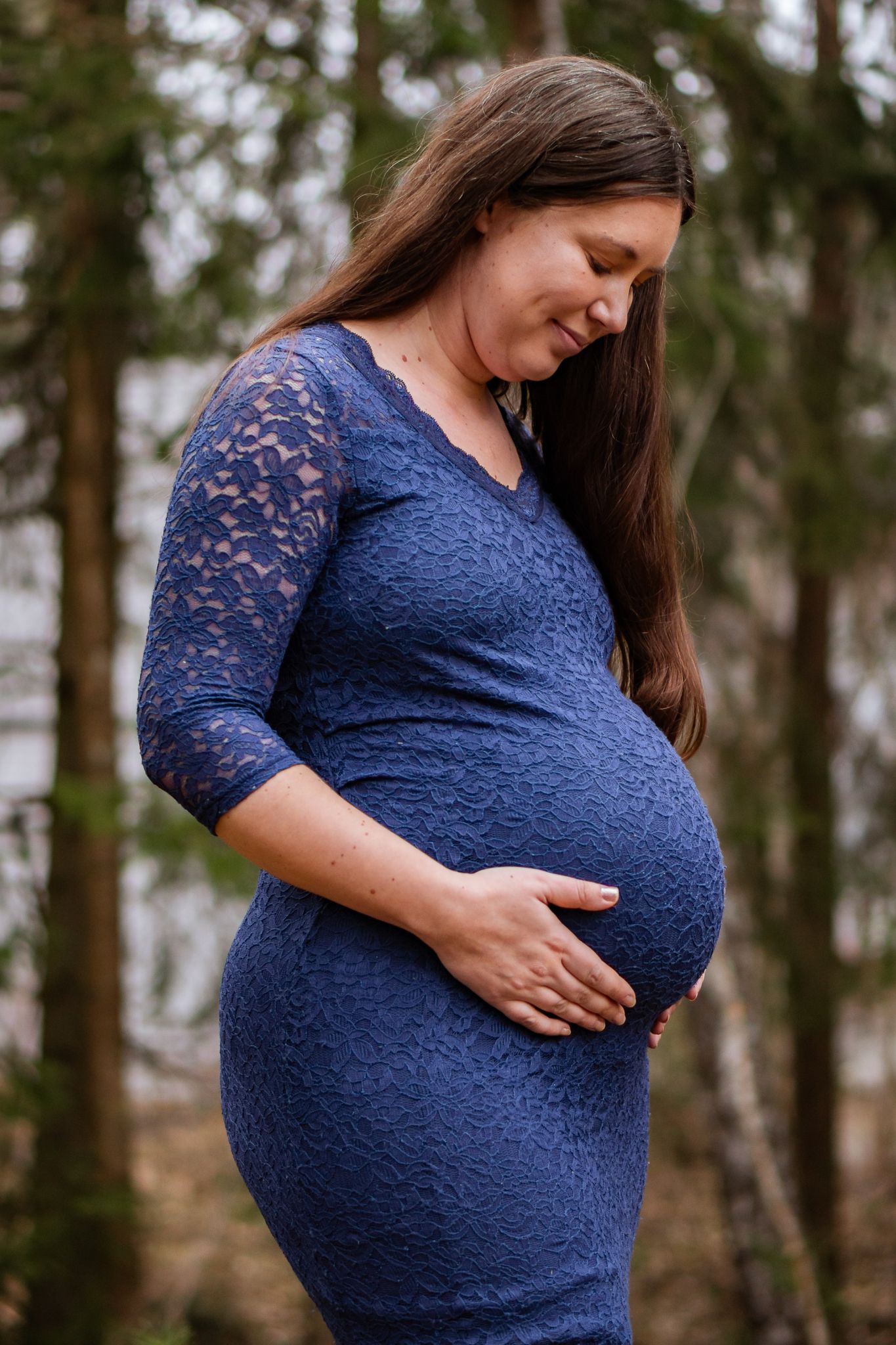 En gravid kvinna i blå spetsklänning tittar ner och ler när hon håller om sin gravida mage i skogen. Gravidfotograf, Umeå, Västerbotten