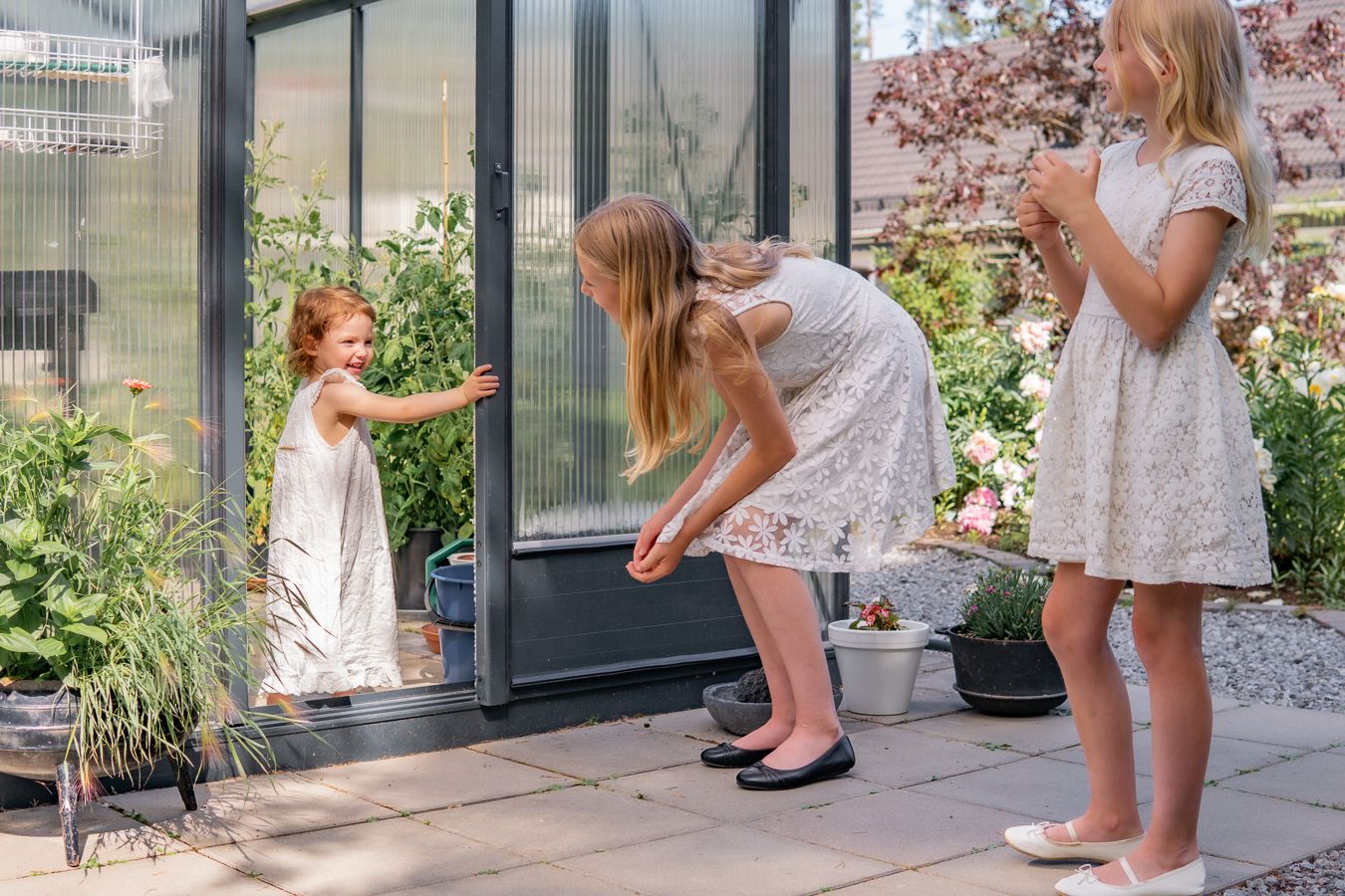 Tre flickor i vita klänningar utforskar ett växthus i trädgården en solig sommardag. Fotograf, Familjefotograf, Umeå, Västerbotten