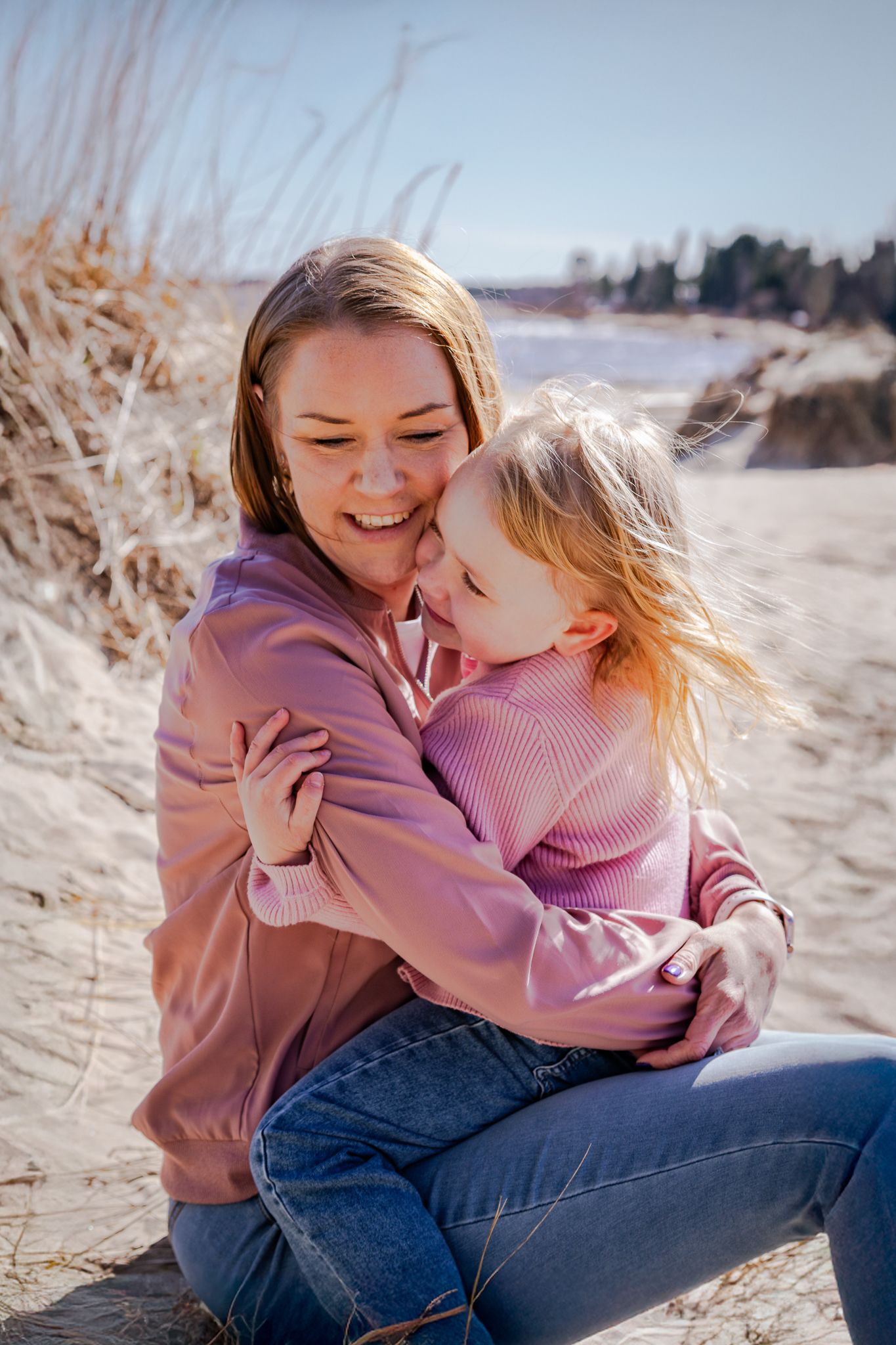 Mamma sitter kärleksfullt med dotter i knät vid sanddynorna på stranden en solig dag. Fotograf, familjefotograf, Umeå