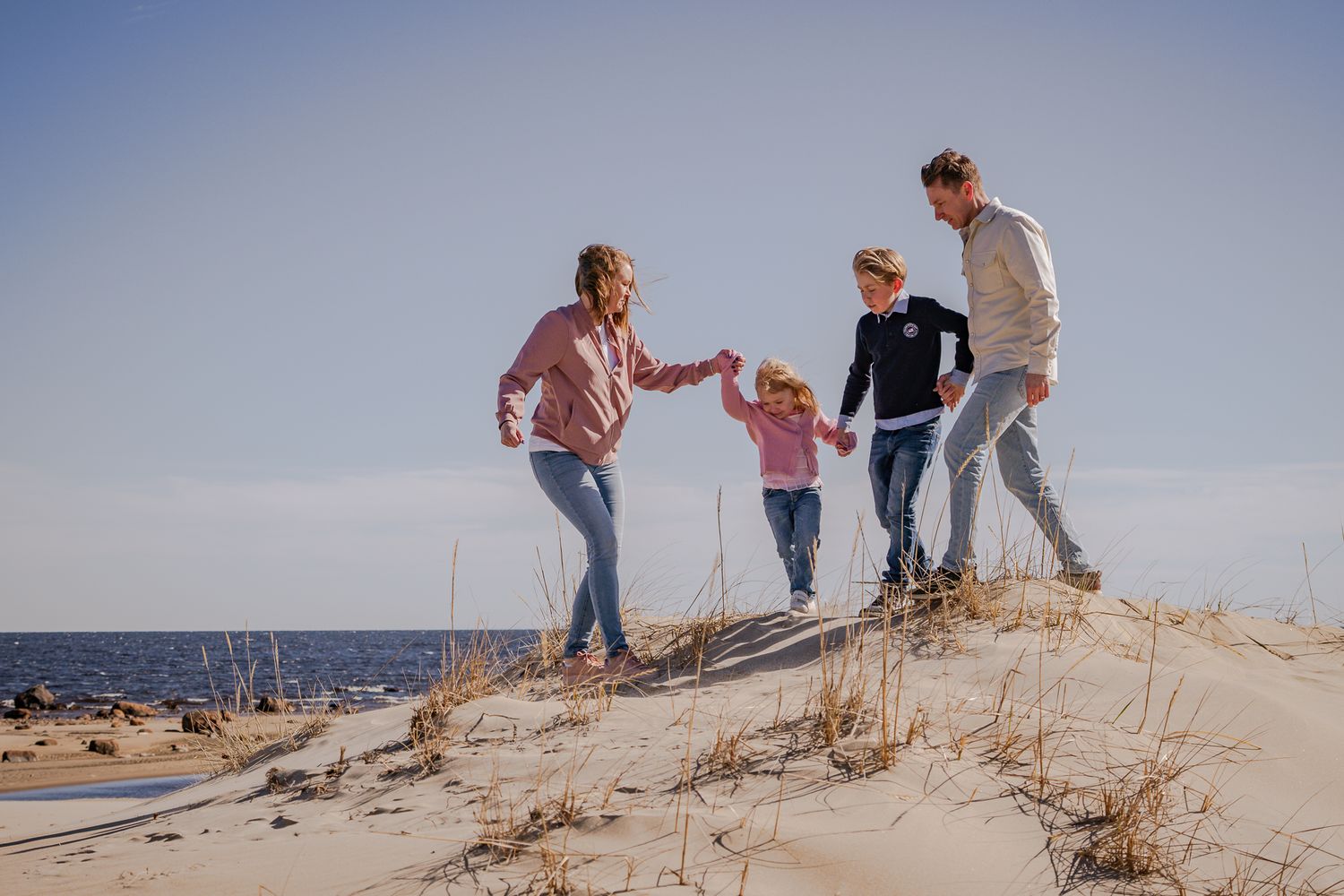 Familj som går hand i hand och leder varandra över sanddynorna på stranden en blåsig solig dag. Fotograf, familjefotograf, Umeå