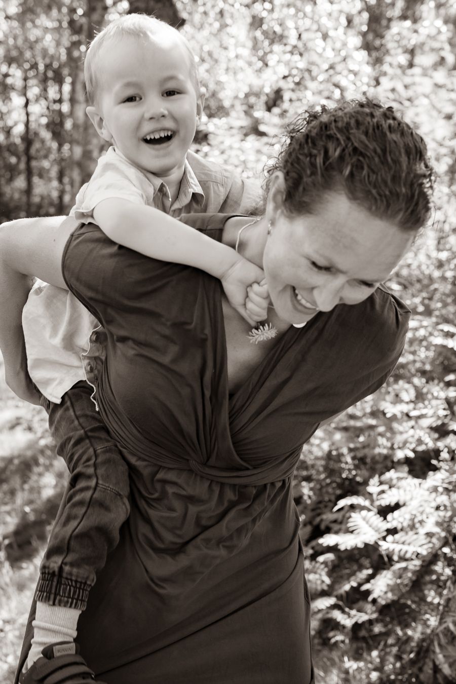 Ett lekfullt ögonblick utomhus då ett barn rider på sin mammas rygg med träd i bakgrunden. Fotograf, Familjefotograf, Umeå